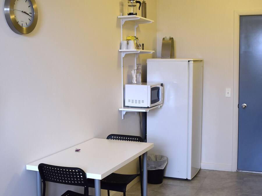 A compact kitchenette with a white refrigerator, a microwave, and a small shelf with kitchen items. There is a white table with two chairs and a clock on the wall.