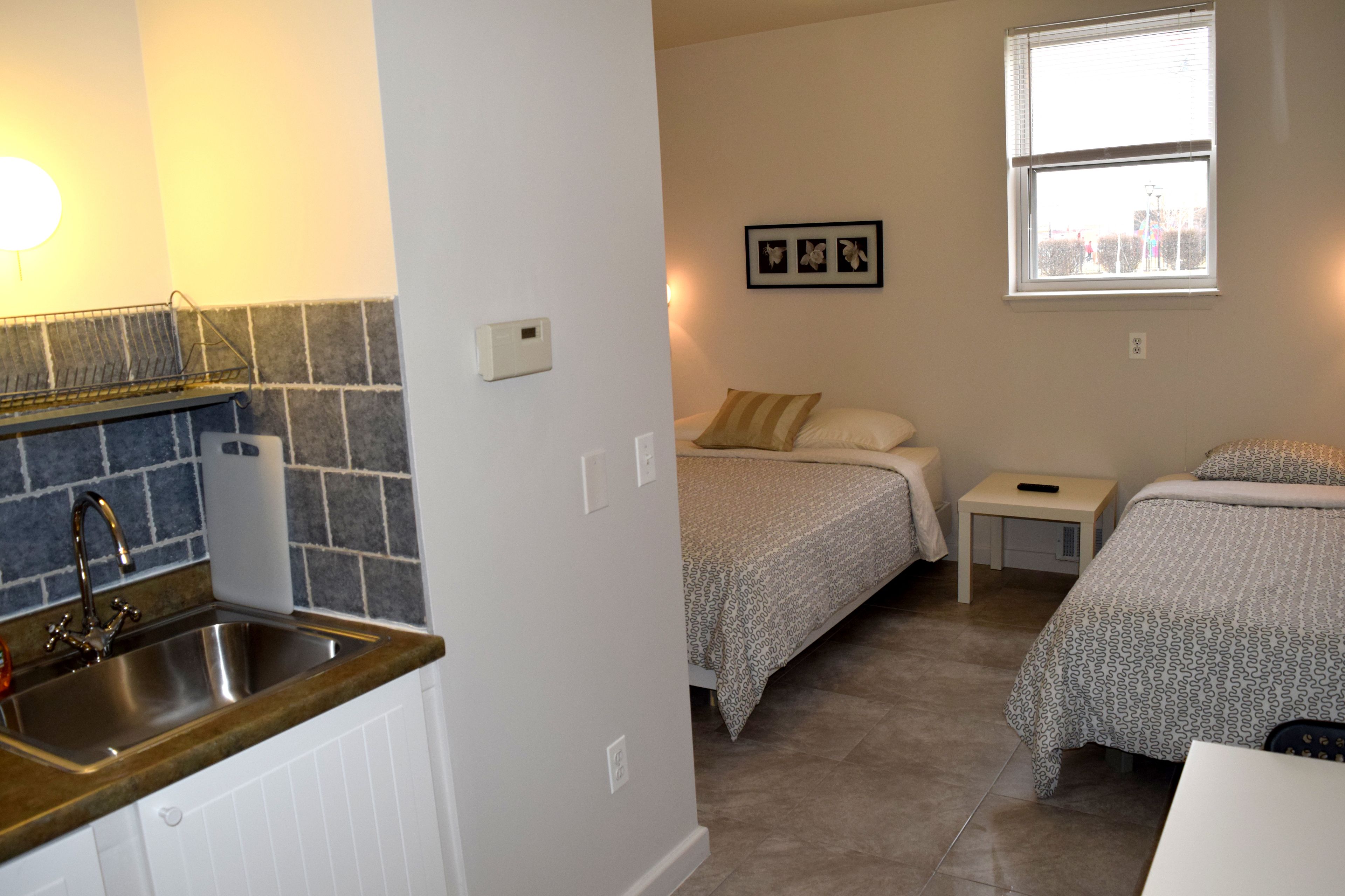A compact hotel room with a kitchenette, a bed with patterned bedding, a bedside table, and a window with a city view.