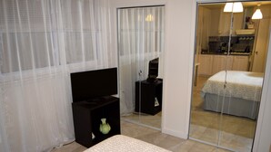A bedroom with a bed, a bedside table, a mirror, and a view of a kitchen through a glass door.