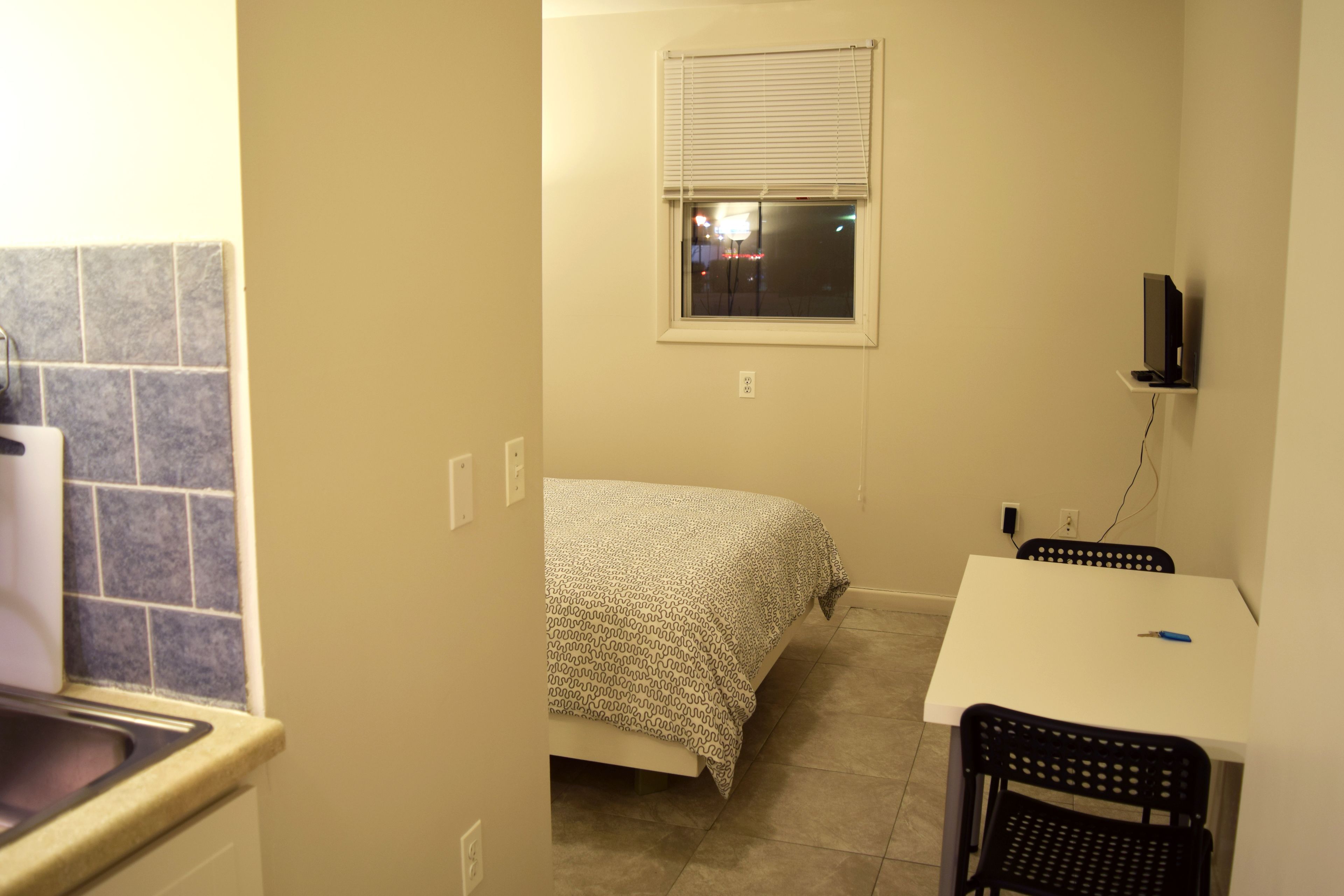 A small room with a bed, a desk, and a chair. There is a window with blinds, a television on a shelf, and a tiled backsplash in the kitchen area.