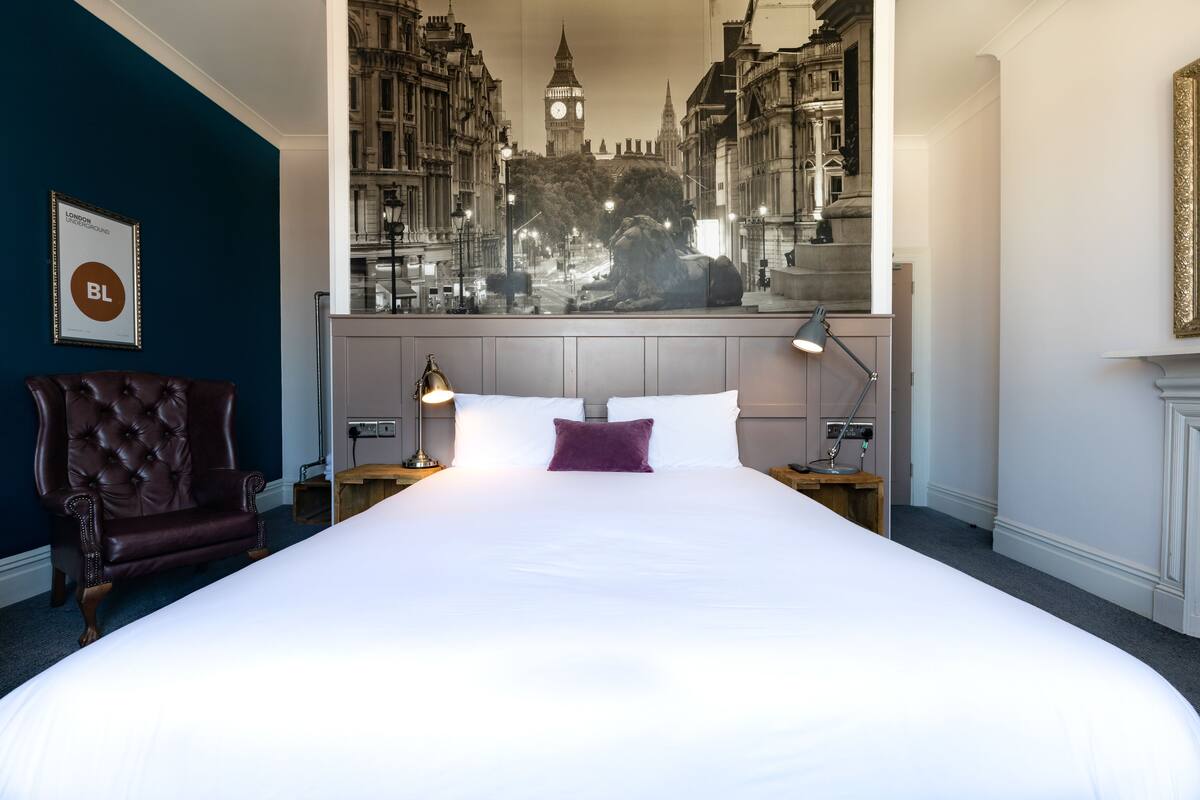 Ein Hotelzimmer mit einem großen Bett, einem Ledersessel, Nachttischlampen und einem Wandgemälde mit Stadtansicht.