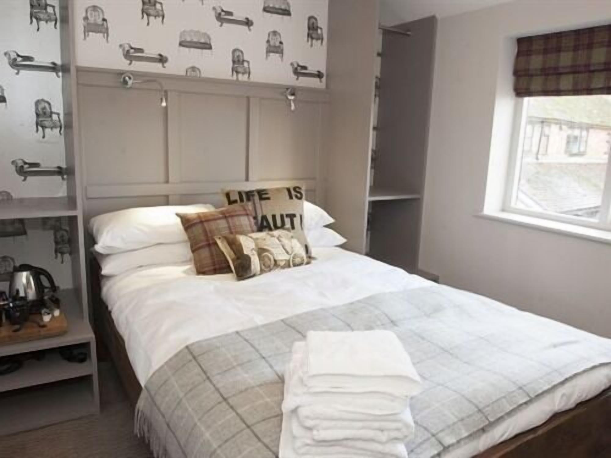 A neatly made bed with a plaid bedspread and pillows, a bedside table with a kettle, and a wall with a pattern of chairs.