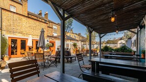 An outdoor patio area with wooden benches and tables, surrounded by brick buildings and potted plants. - The Station Hotel (London)