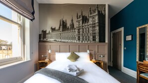 A hotel room with a large bed, a nightstand with a lamp, a mirror, and a view of a building through the window. - The Station Hotel (London)