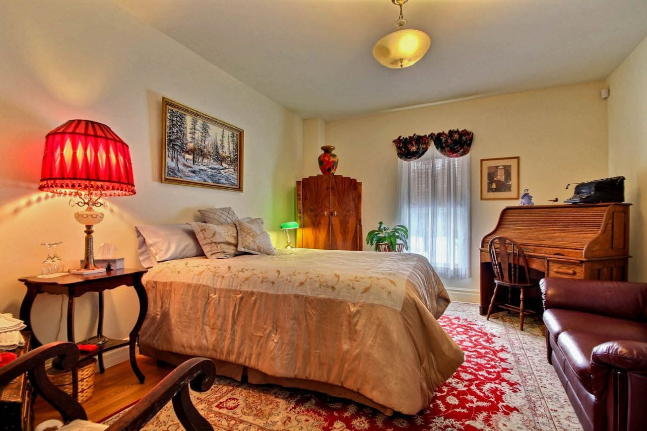 A cozy bedroom with a bed, a wooden dresser, a desk with a piano, and a red lamp.