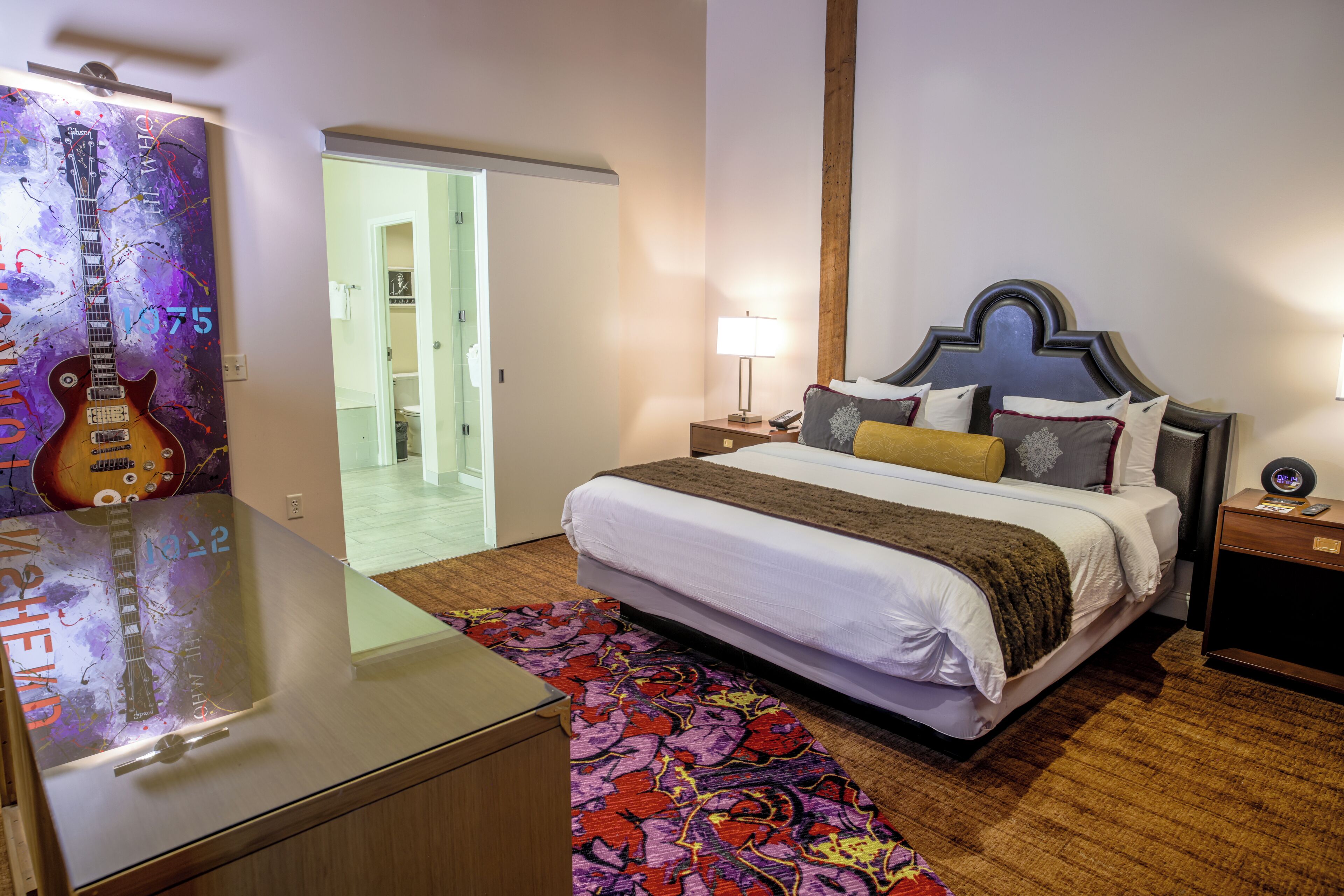 A hotel room with a bed, bedside tables, a lamp, a guitar, and a colorful rug.