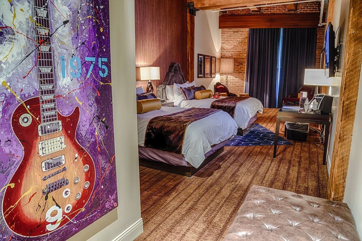 A hotel room with a large guitar mural, two beds, a desk, and a brick wall.