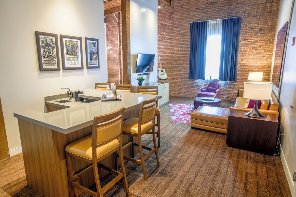 A modern hotel room with a kitchen area, a seating area with a sofa and chairs, and a large window with curtains.