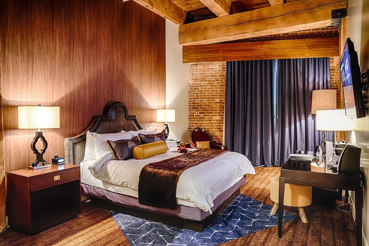 A hotel room with a wooden ceiling, a brick wall, a large bed with multiple pillows, a desk with a computer, and a lamp.