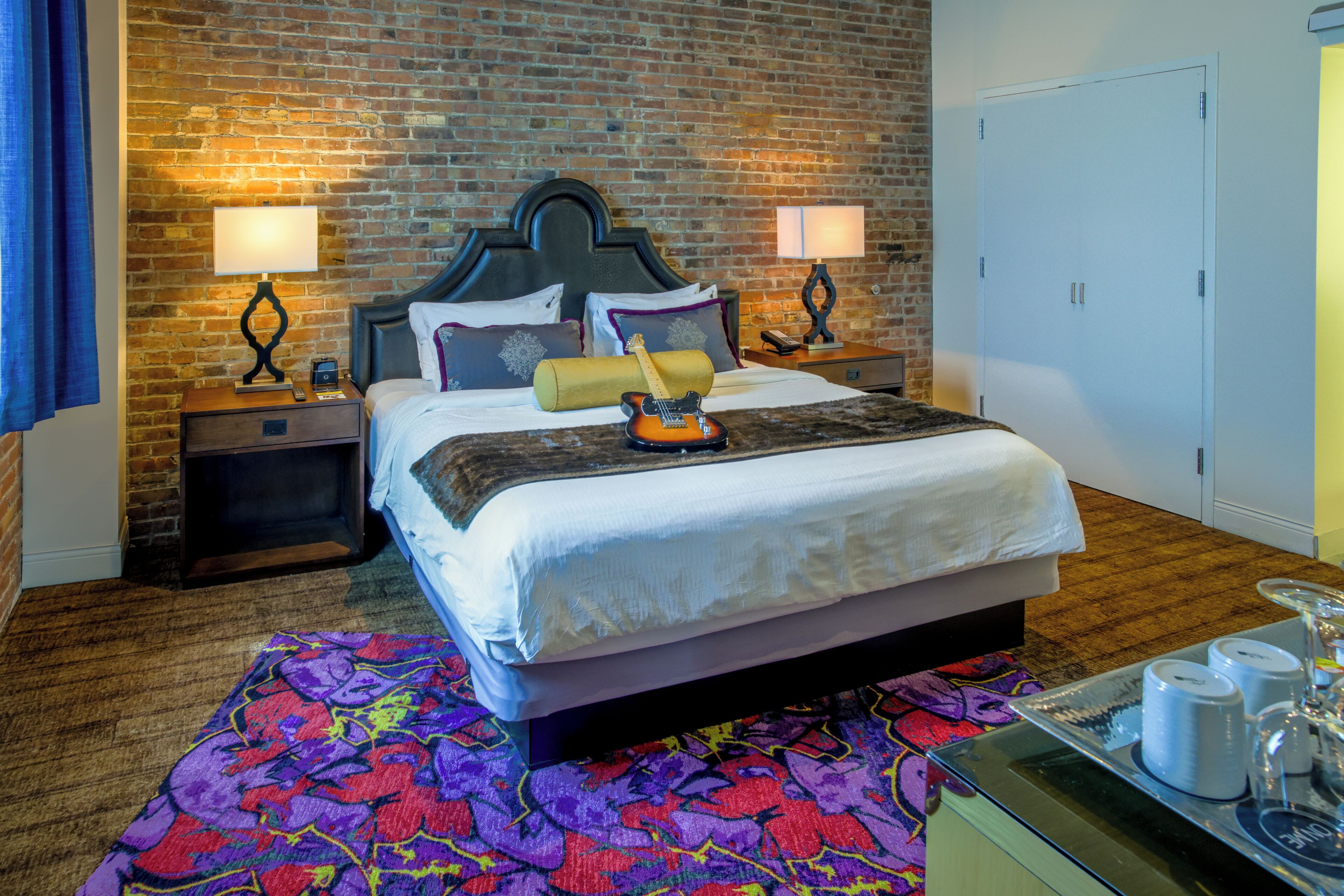 A hotel room with a brick wall, a bed with a wooden headboard, two bedside tables with lamps, a colorful rug, and a tray with glasses and a bottle.