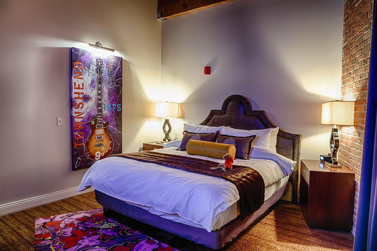 A bedroom with a bed, two bedside tables, a lamp, a colorful rug, and a large abstract painting on the wall.