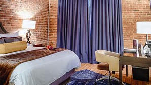 A room with exposed wooden beams, a brick wall, a bed with a white comforter, a blue patterned rug, a desk with a chair, and a large window with dark curtains. - Hard Rock Hotel & Casino Sioux City (Sioux City)