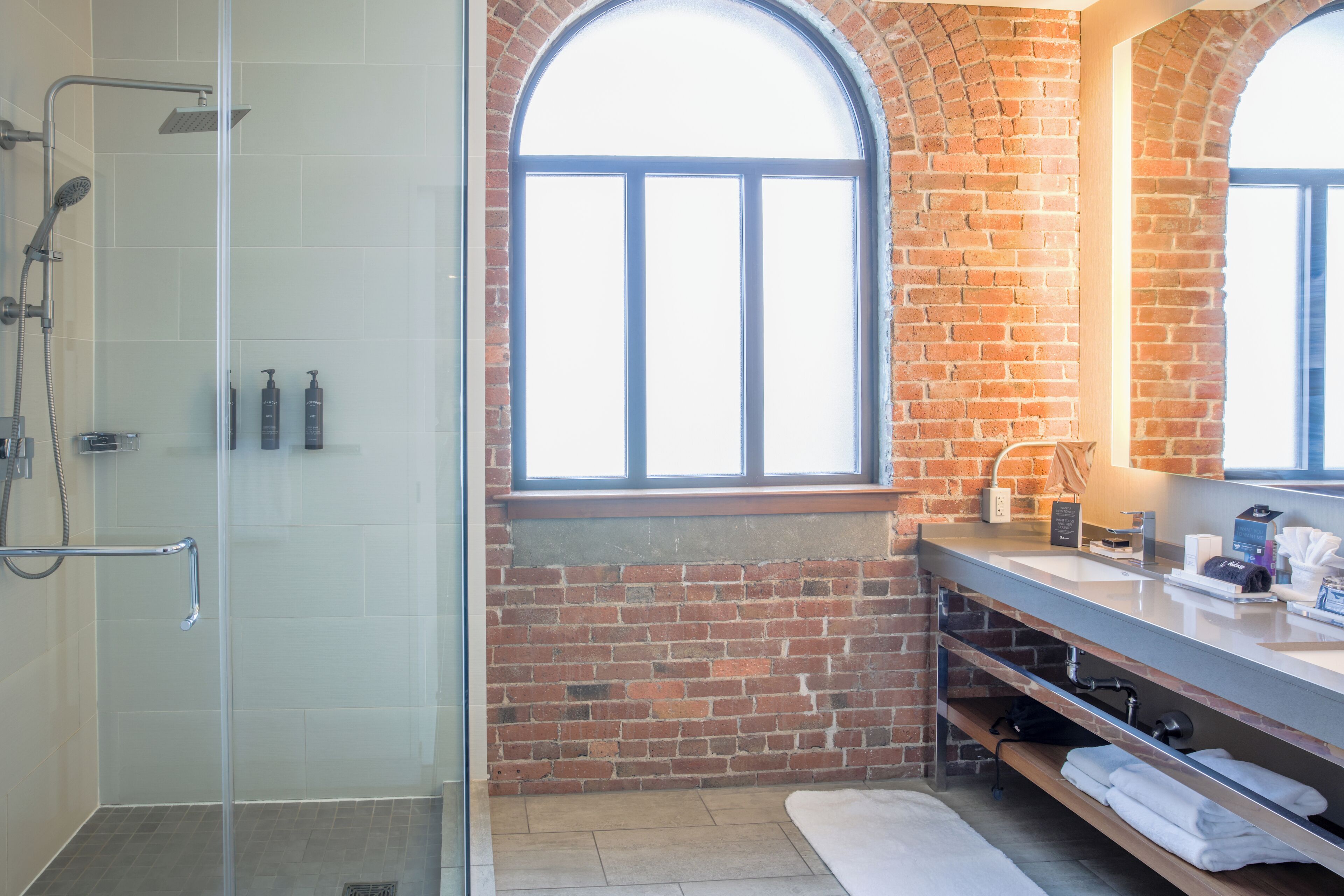 A modern bathroom with a glass shower enclosure, a brick wall, and a large arched window.