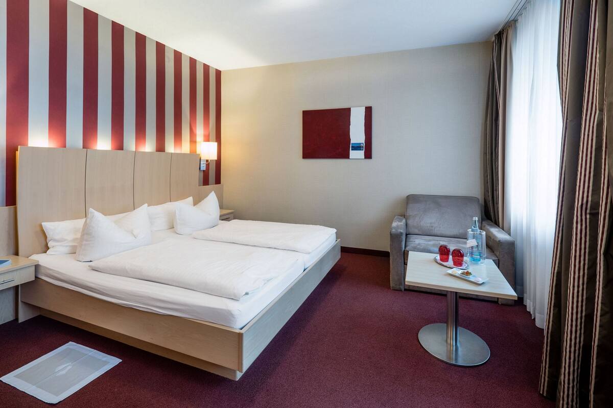 Ein Hotelzimmer mit einem großen Bett, einem Nachttisch mit Lampe, einem kleinen Tisch mit Flasche und Gläsern sowie einem Sessel.