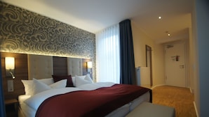 Een hotelkamer met een groot bed, een hoofdbord met een patroon, een nachtkastje met een lamp, en een deur naar een andere kamer.