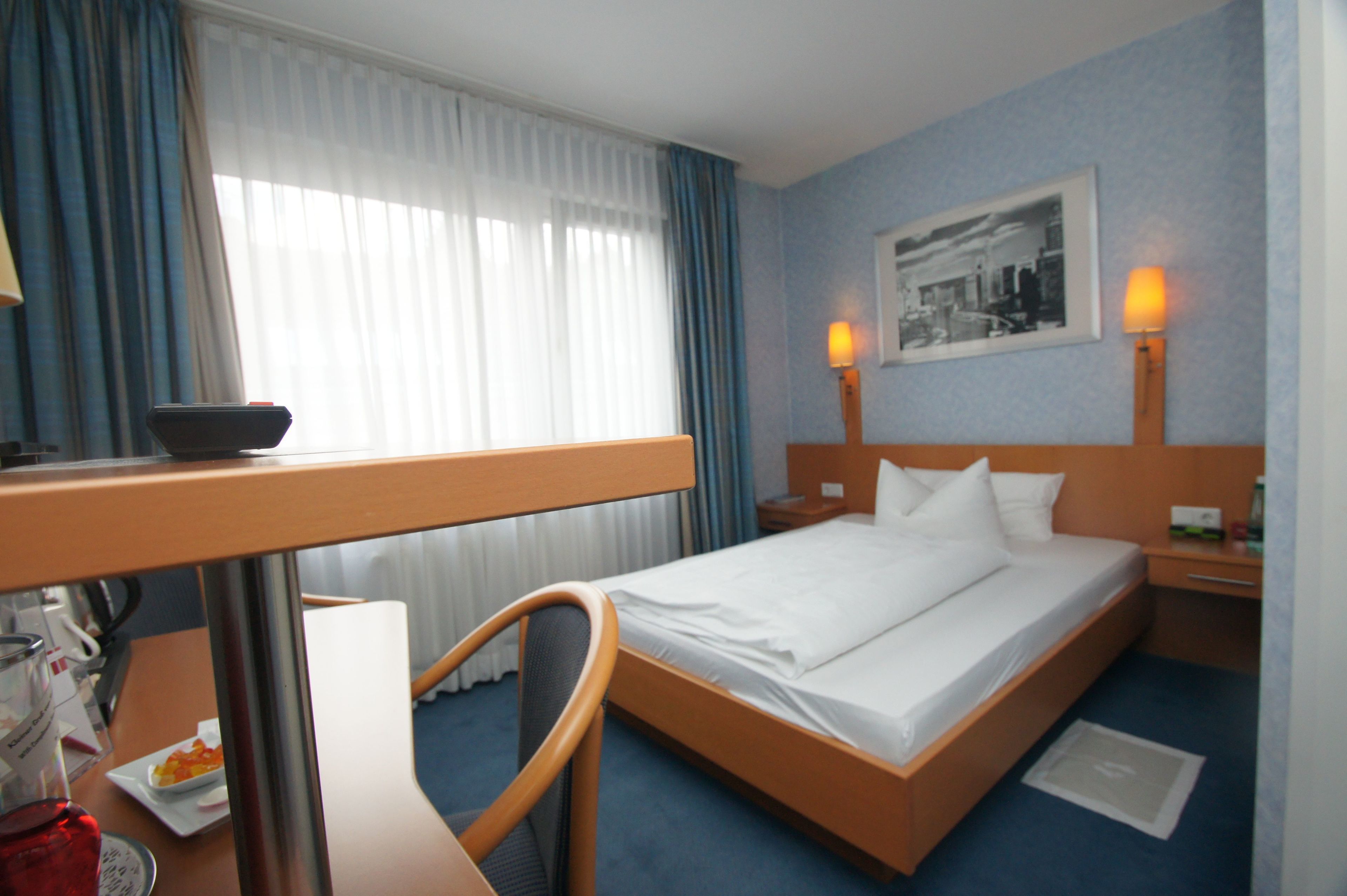 A hotel room with a bed, a desk with a chair, a lamp, and a framed picture on the wall.