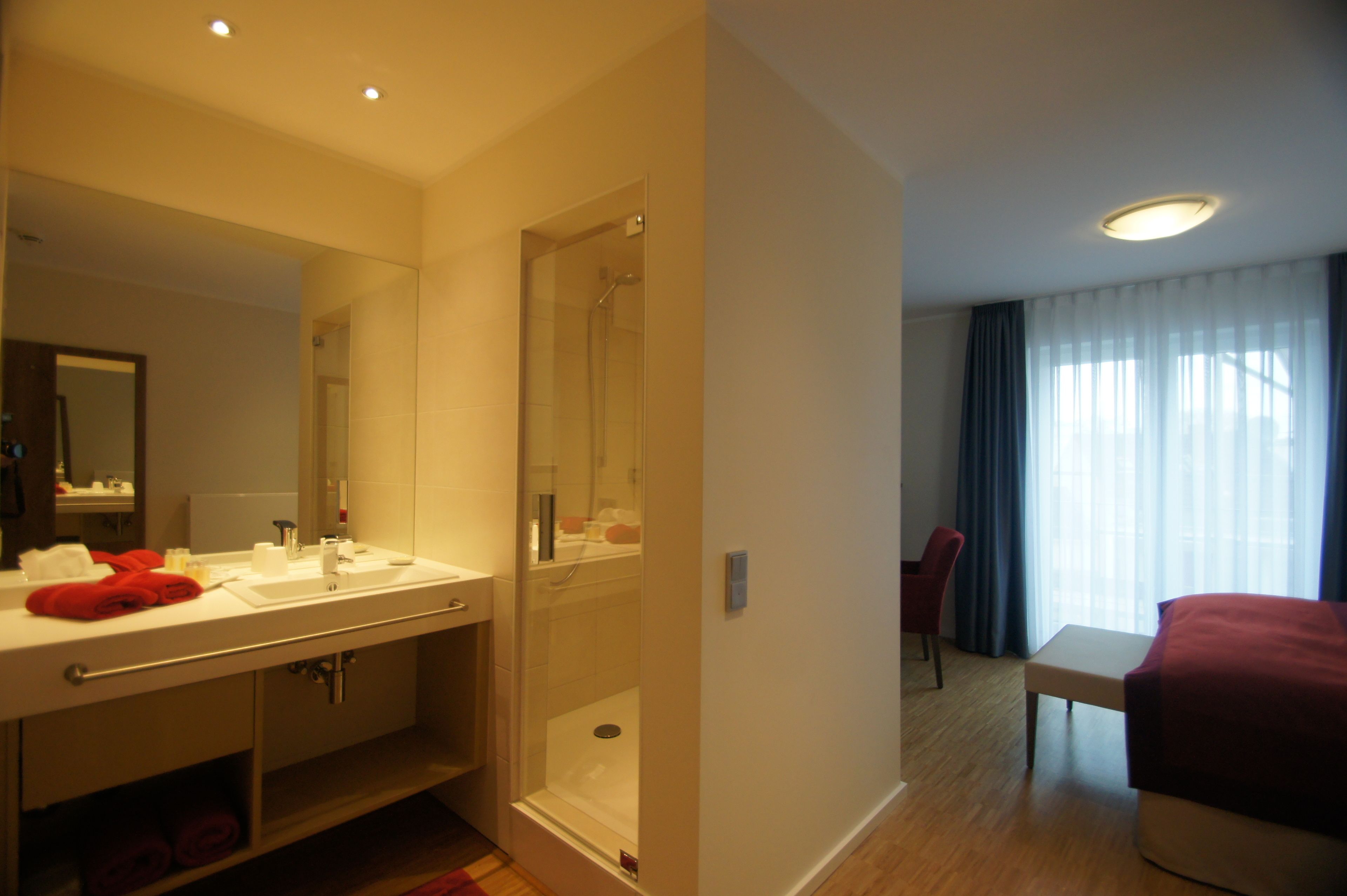 A hotel room with a bathroom featuring a sink, mirror, and shower. There is a bed and a chair in the room.