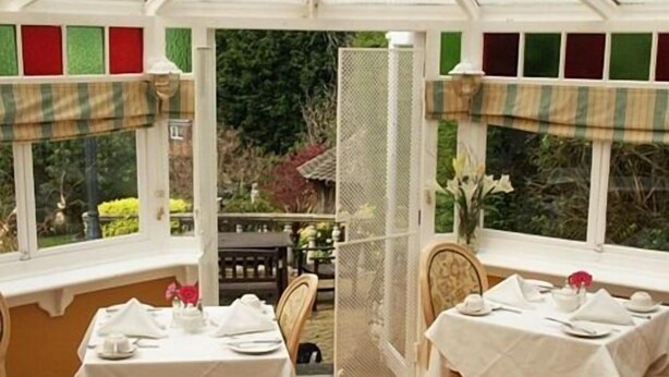 Un invernadero con dos mesas de comedor preparadas para una comida, rodeado de ventanas con paneles decorativos.