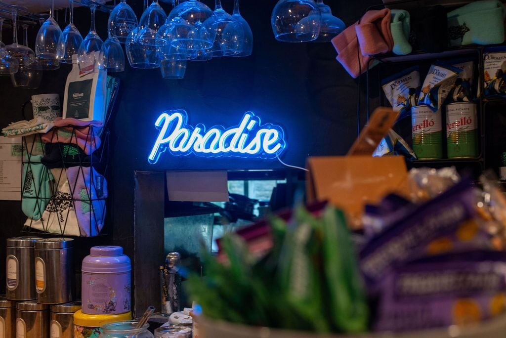 A bar area with a neon sign reading "Paradise," various bottles, and a person holding a menu.