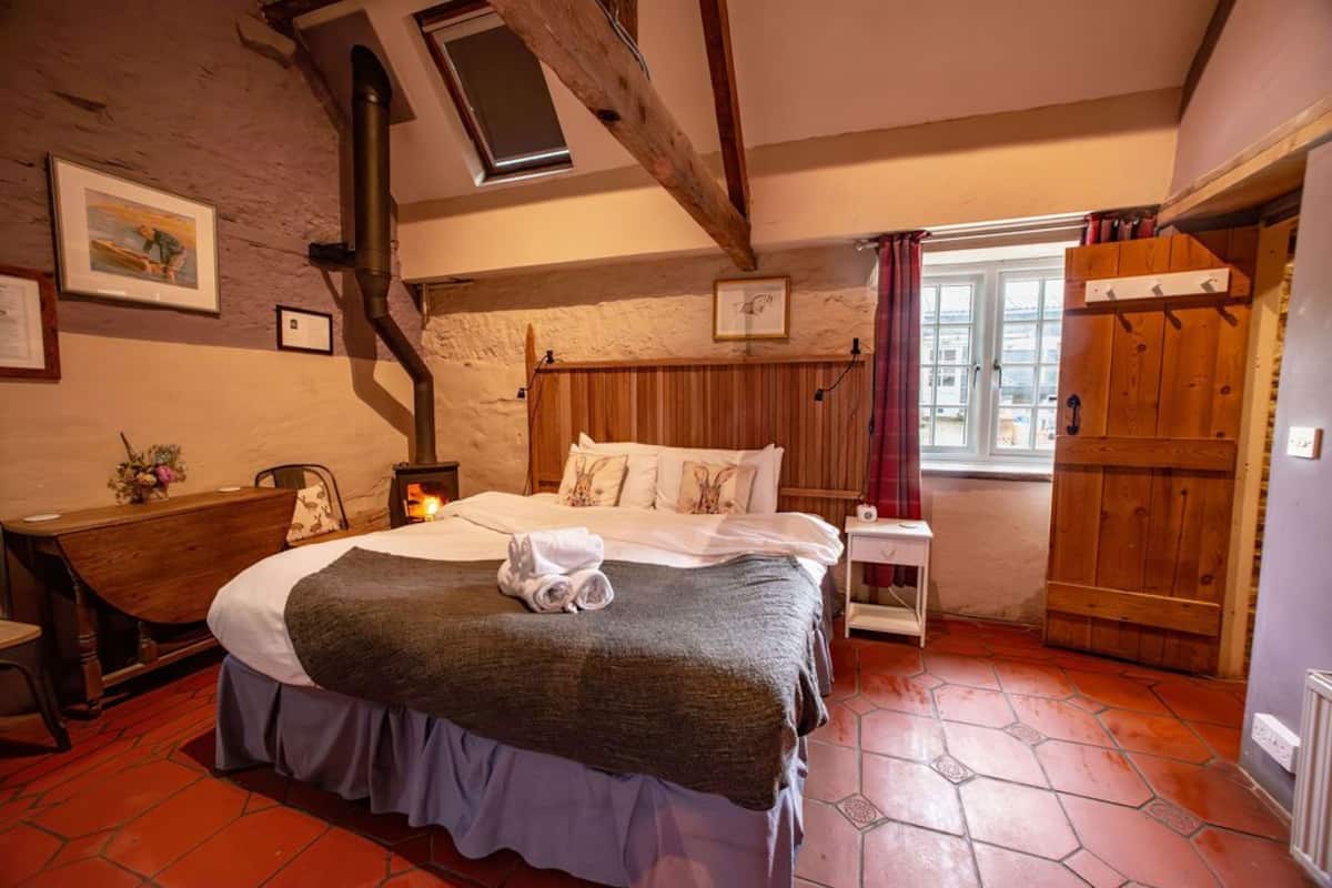 A cozy bedroom with a wooden bed, a fireplace, a desk, and a window with curtains.