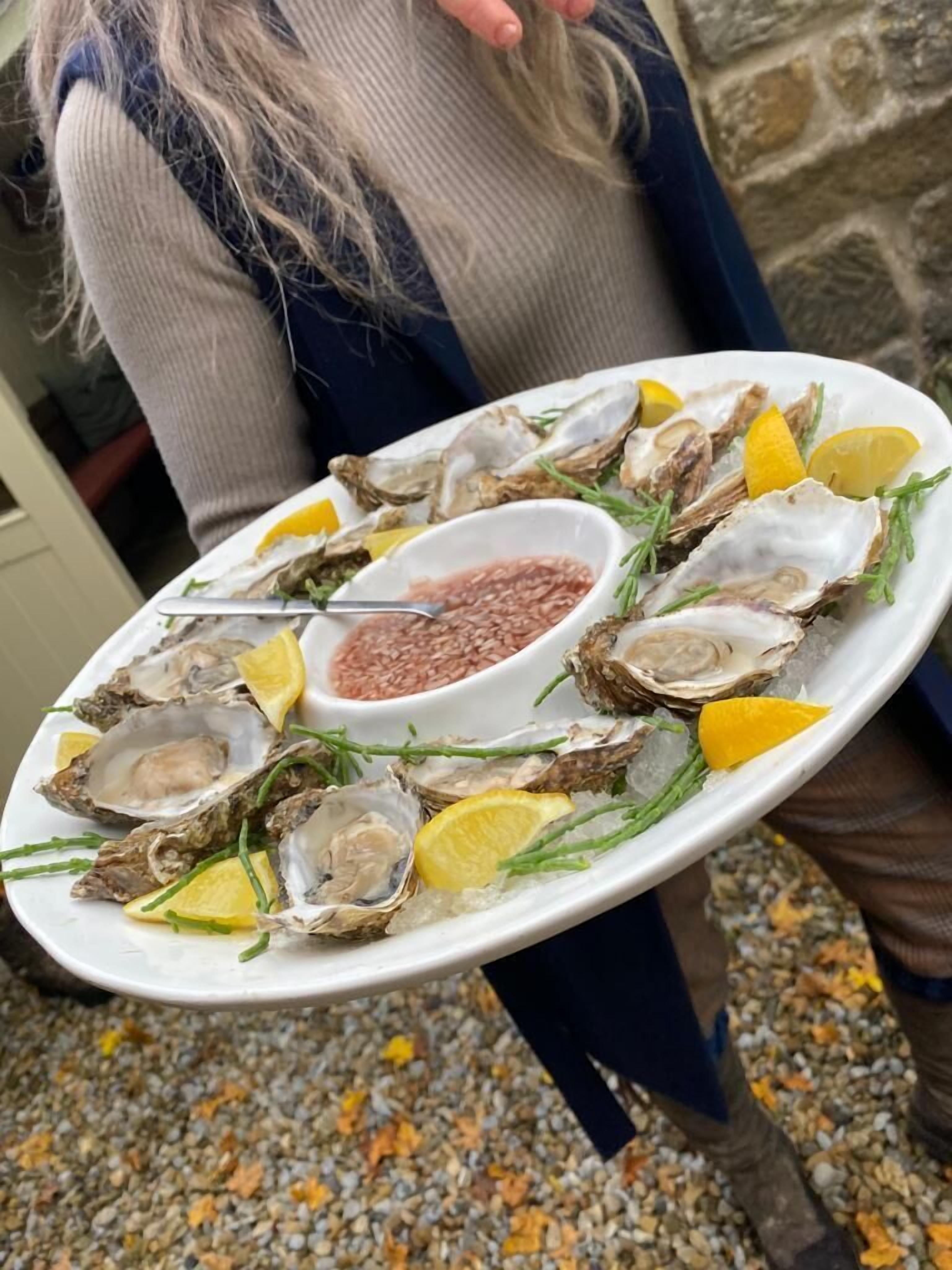 A person holding a plate of oysters with lemon slices and a small bowl of sauce.