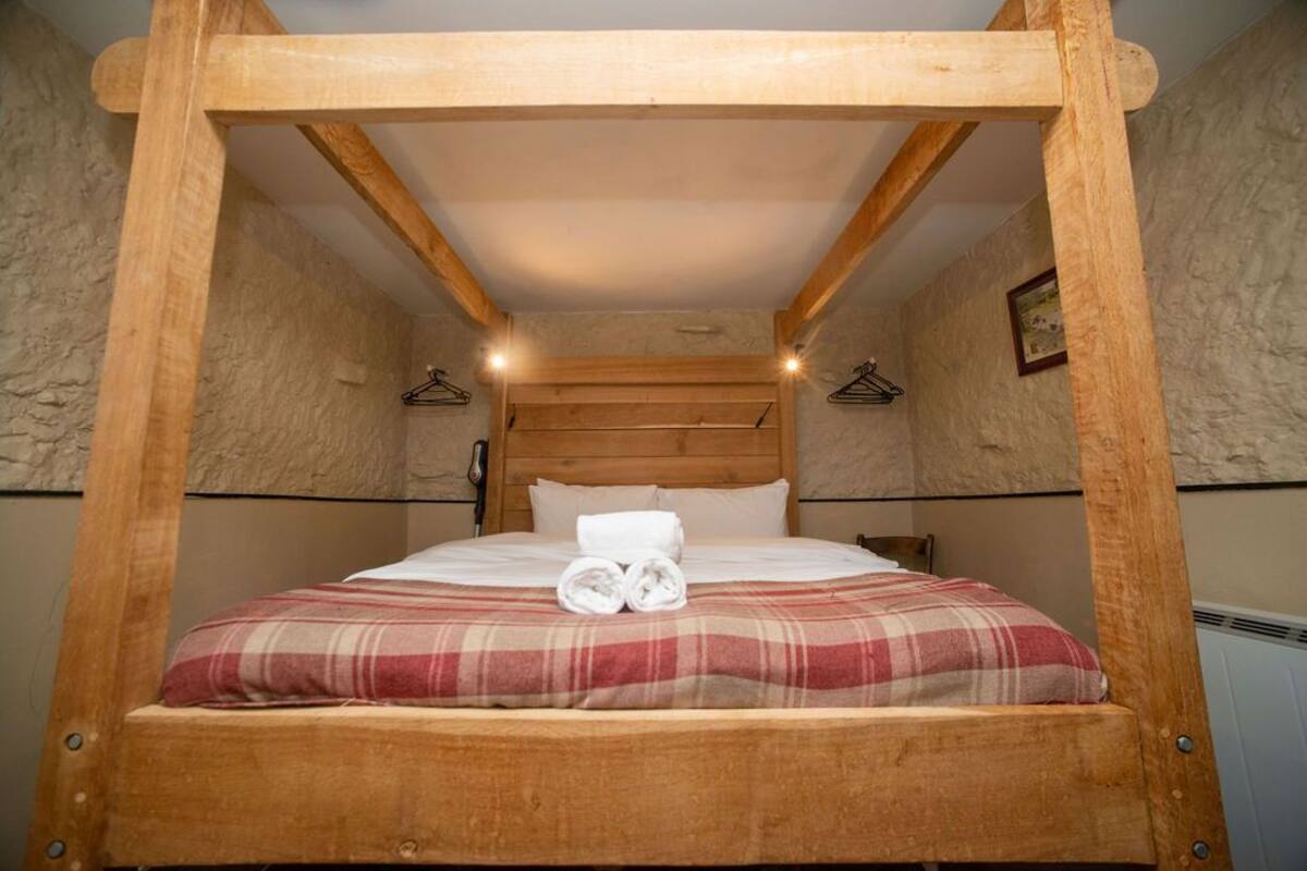 A wooden four-poster bed with a plaid bedspread and rolled towels, set in a room with stone walls and wooden beams.
