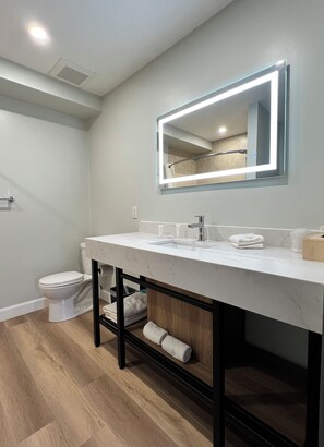 A modern bathroom with a white vanity, a large mirror with LED lighting, a toilet, and a wooden floor. - Wellington Inn (Bracebridge)