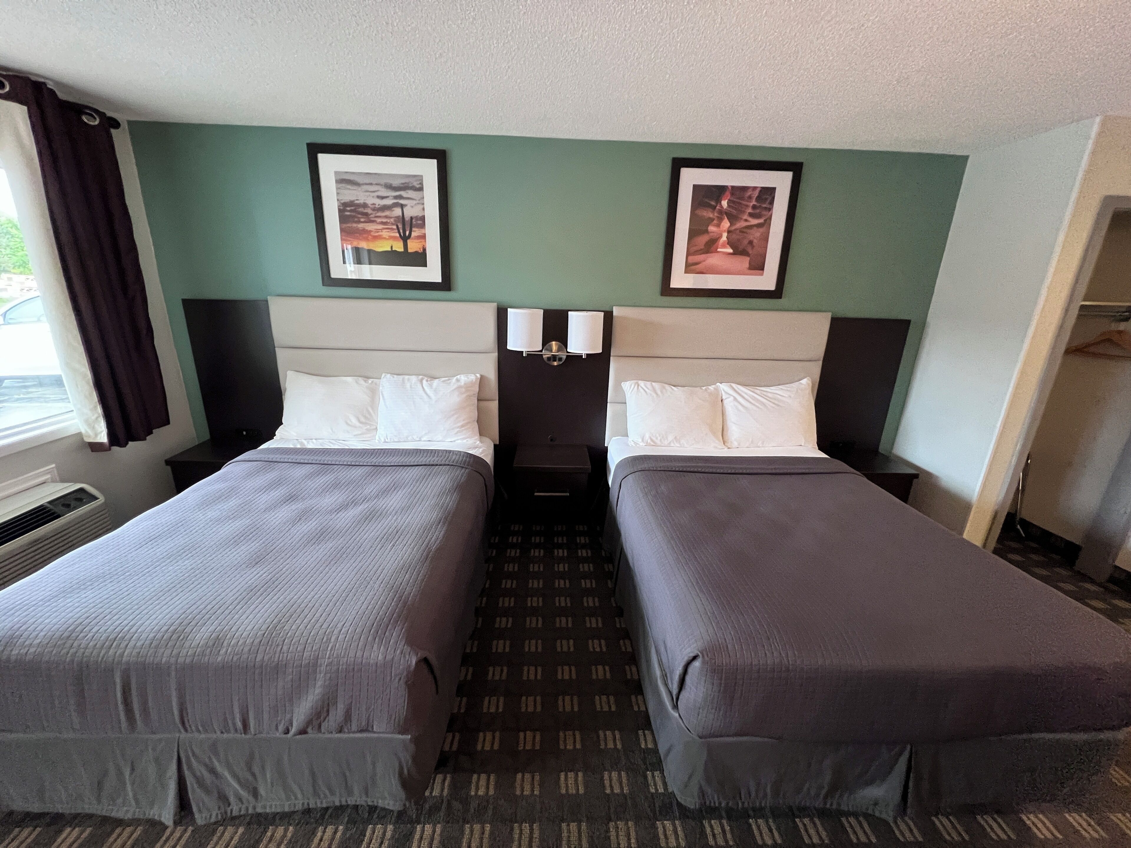 A hotel room with two beds, a window with curtains, a wall-mounted TV, and framed pictures above the beds.