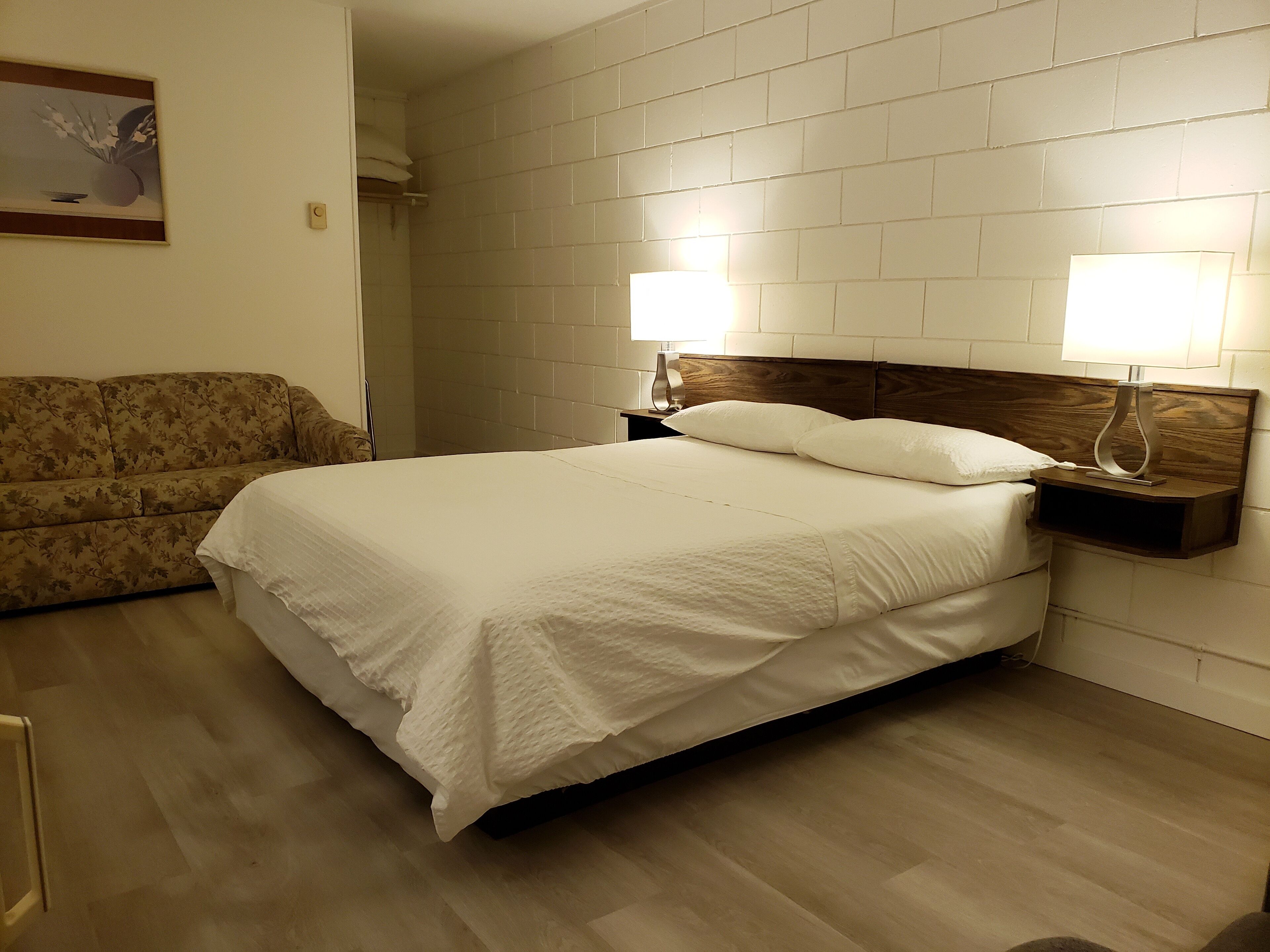 A hotel room with a bed, a nightstand with a lamp, a wooden headboard, a sofa, and a painting on the wall.