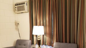 Ein Zimmer mit zwei grauen Sesseln, einem Holztisch, einer Lampe, einem Streifenmuster-Vorhang und einer Wand-Klimaanlage.