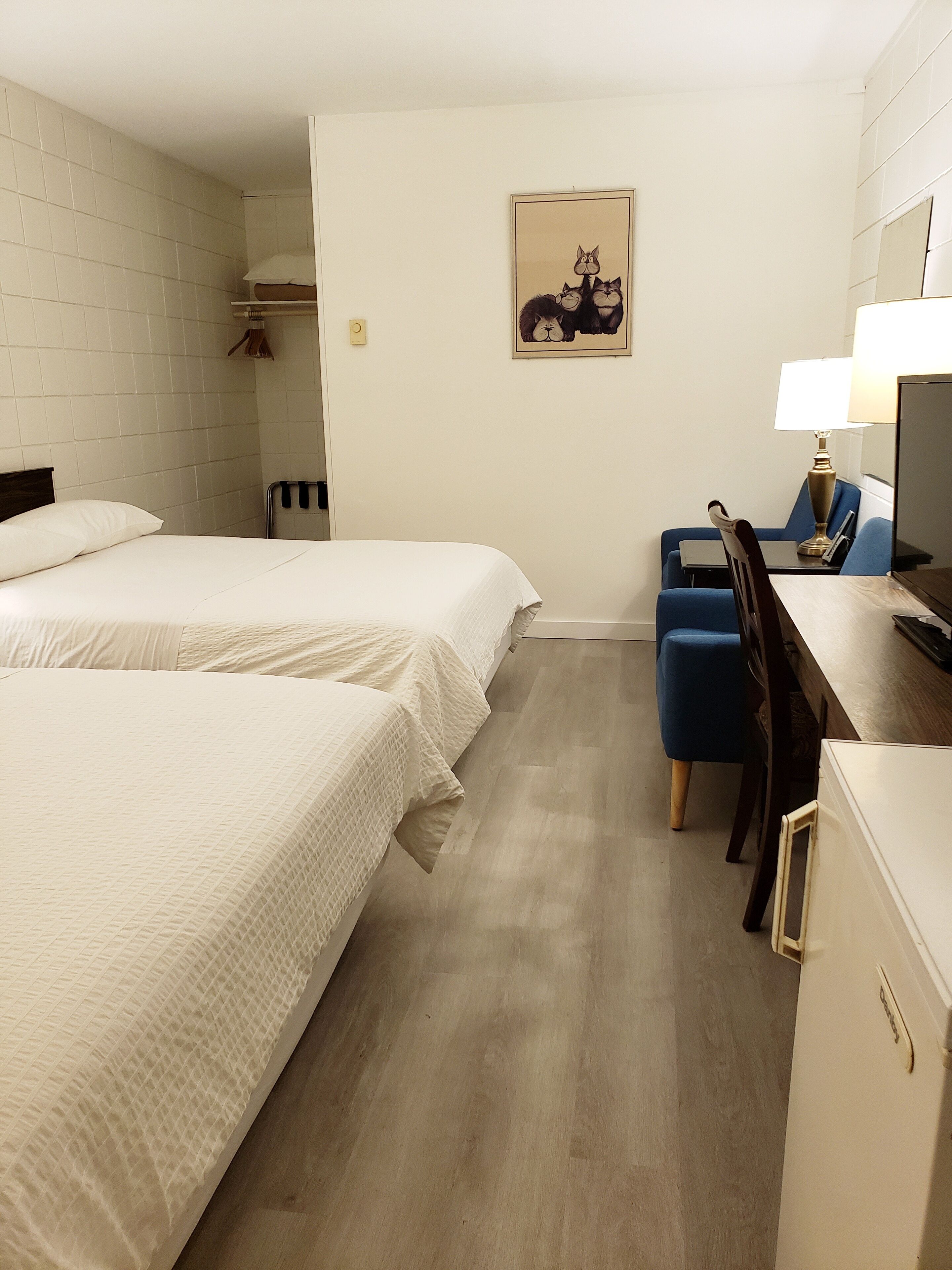 A hotel room with two beds, a desk with a chair, a lamp, and a painting on the wall.