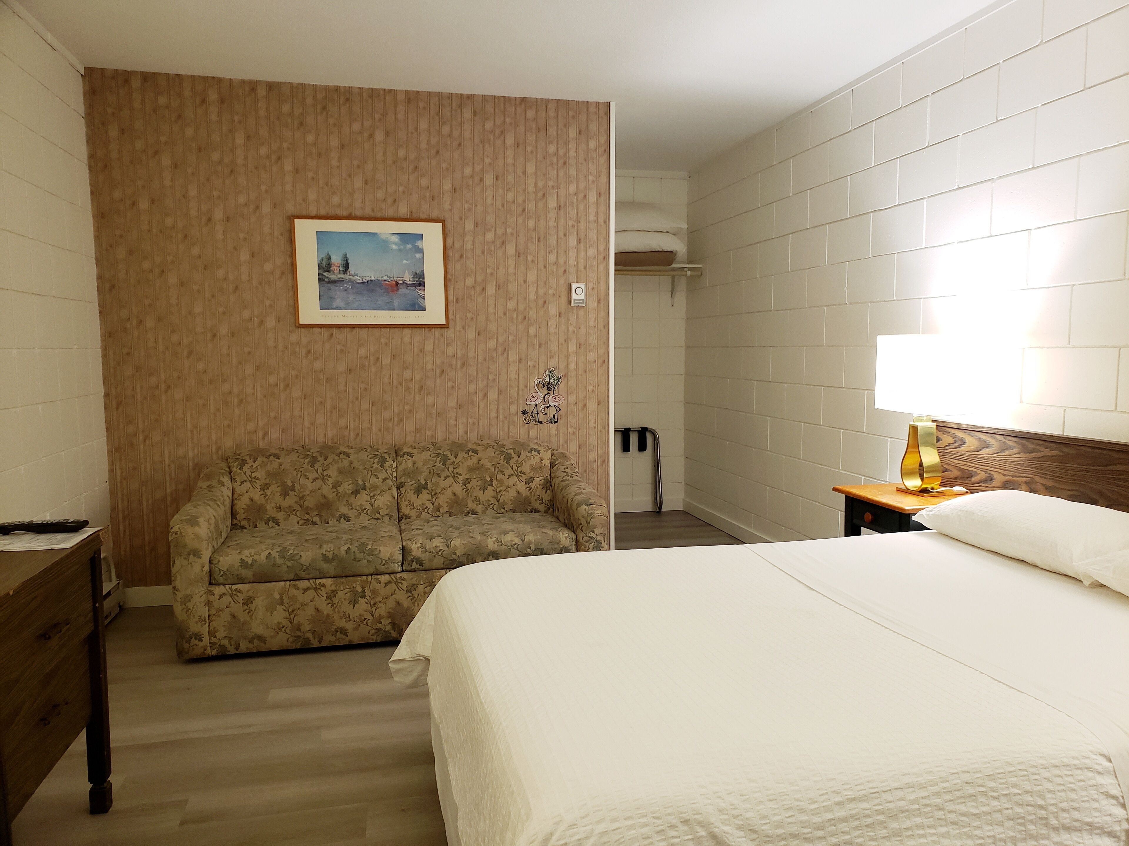 A hotel room with a bed, a sofa, a framed picture on the wall, a bedside table with a lamp, and a wooden floor.