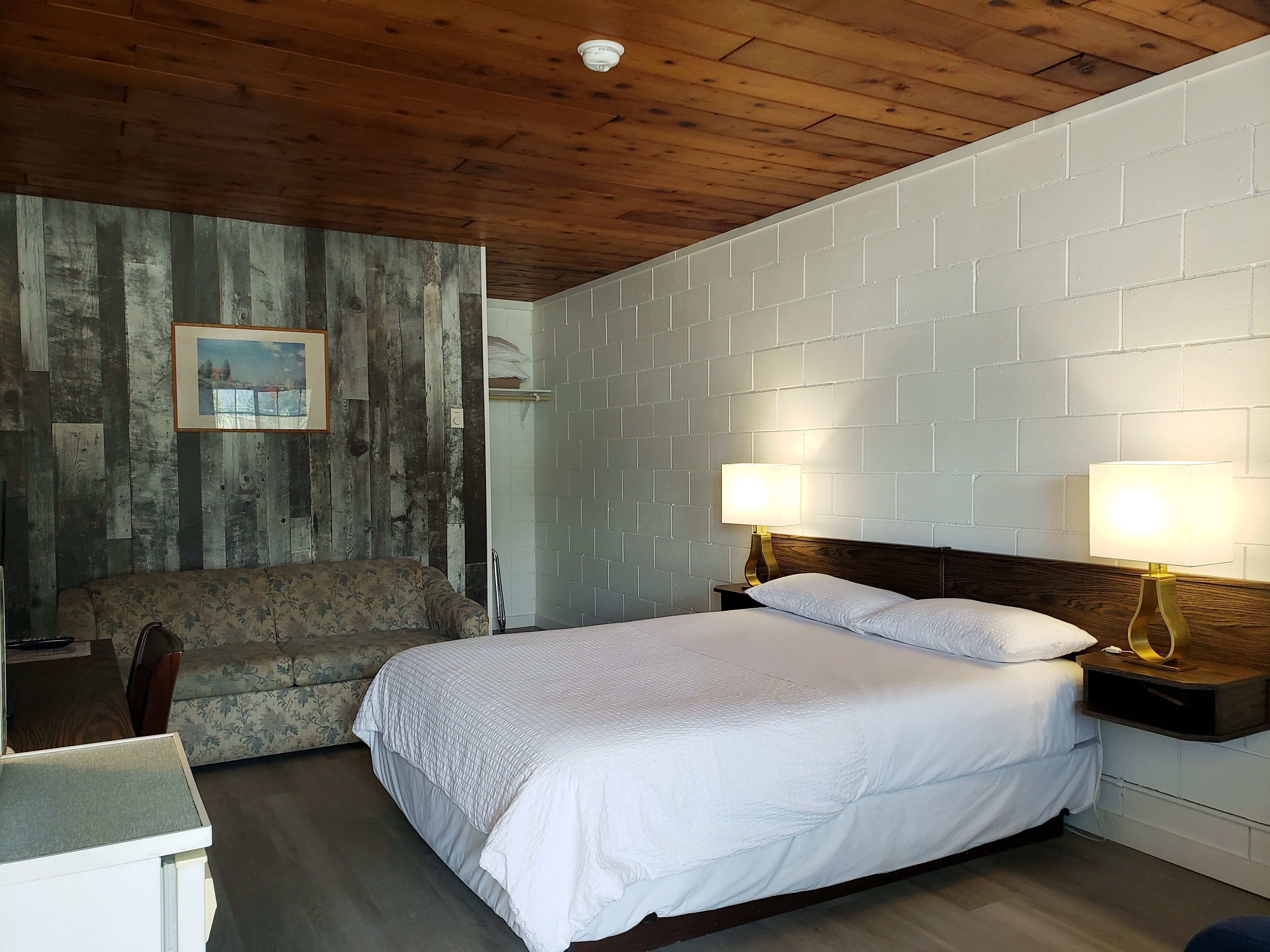 A bedroom with a wooden ceiling, a white brick wall, a bed with white bedding, two bedside lamps, and a framed picture on the wall.
