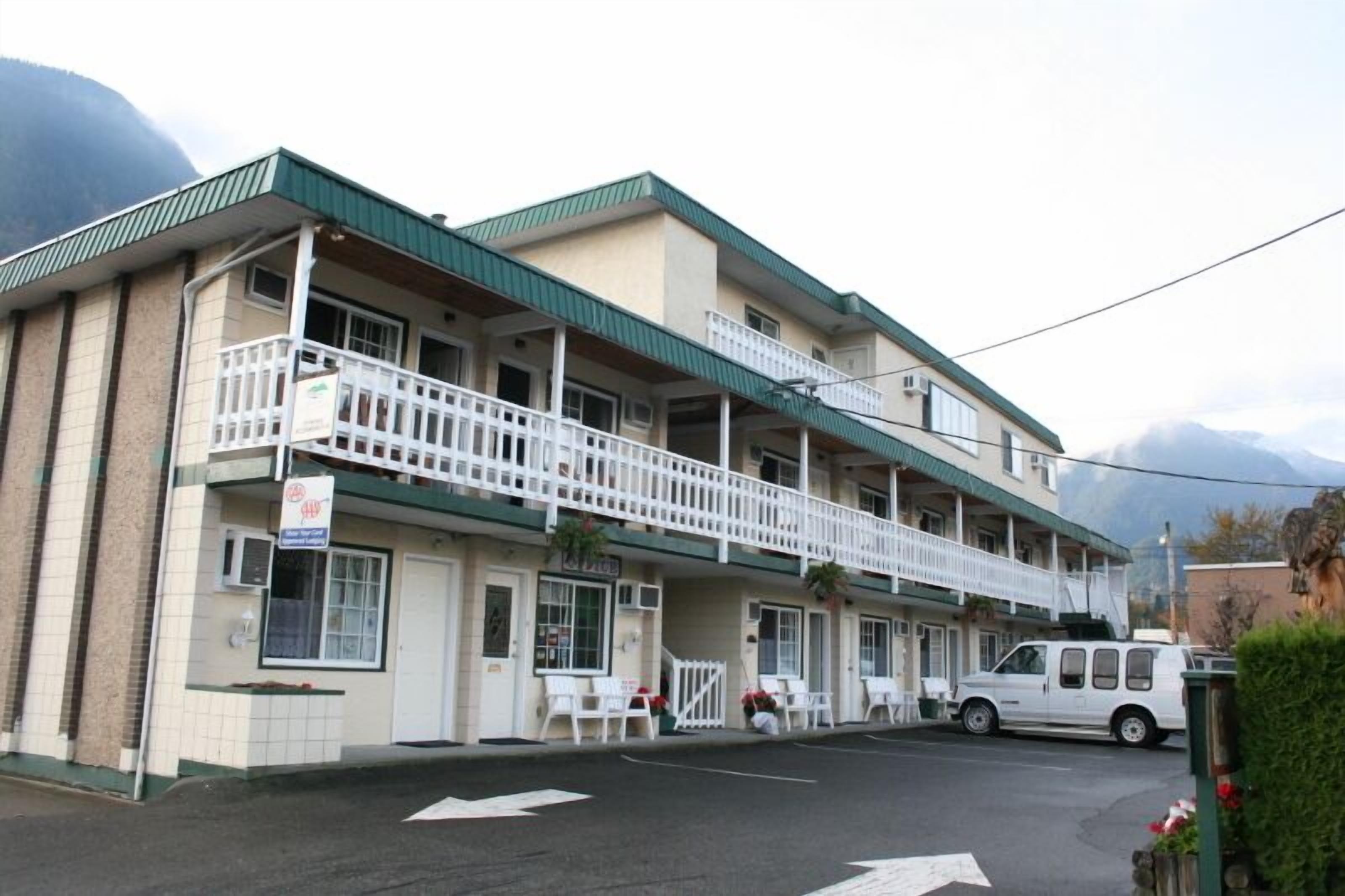 A two-story motel with a green roof, a balcony on the upper floor, and a white van parked in front.