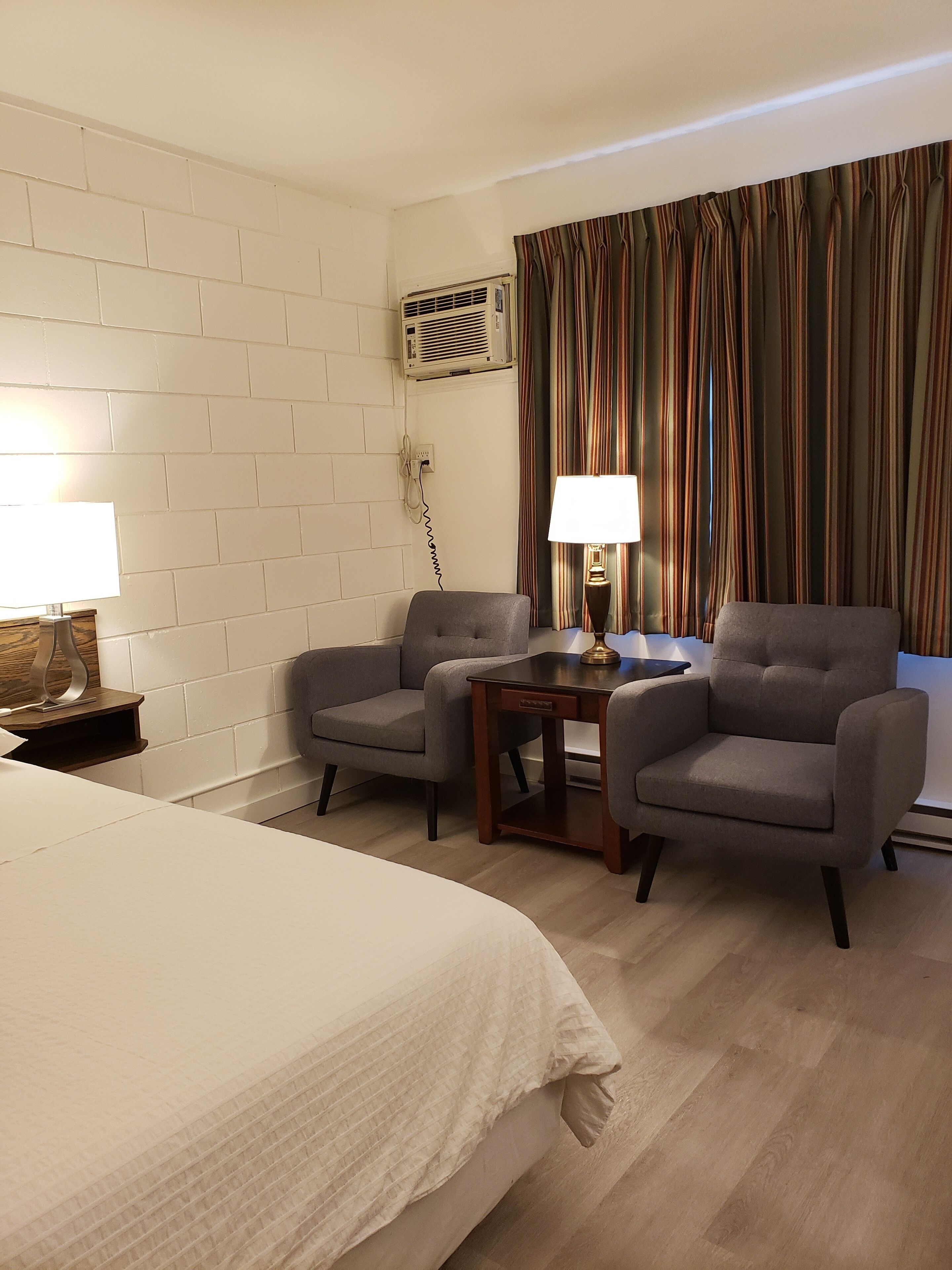Habitación de hotel con una cama grande, dos sillones, una mesita con una lámpara, aire acondicionado de pared y una ventana con cortinas a rayas.