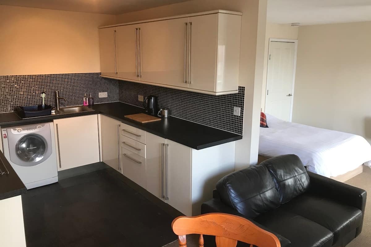 A compact hotel room with a kitchenette, a washing machine, a dining area, a black leather sofa, and a bed.