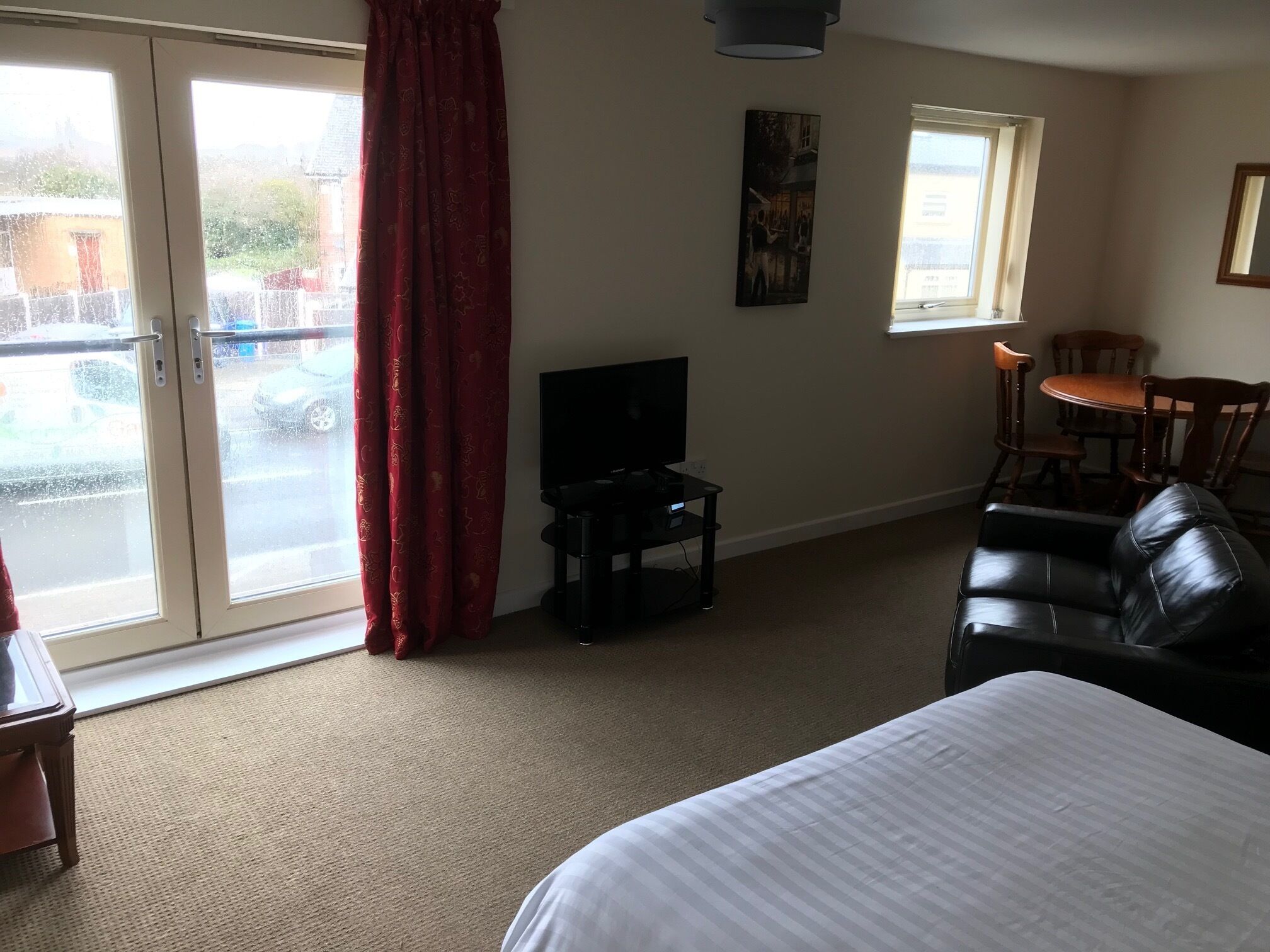 A hotel room with a bed, a TV, a dining table with chairs, and a view through the glass doors.