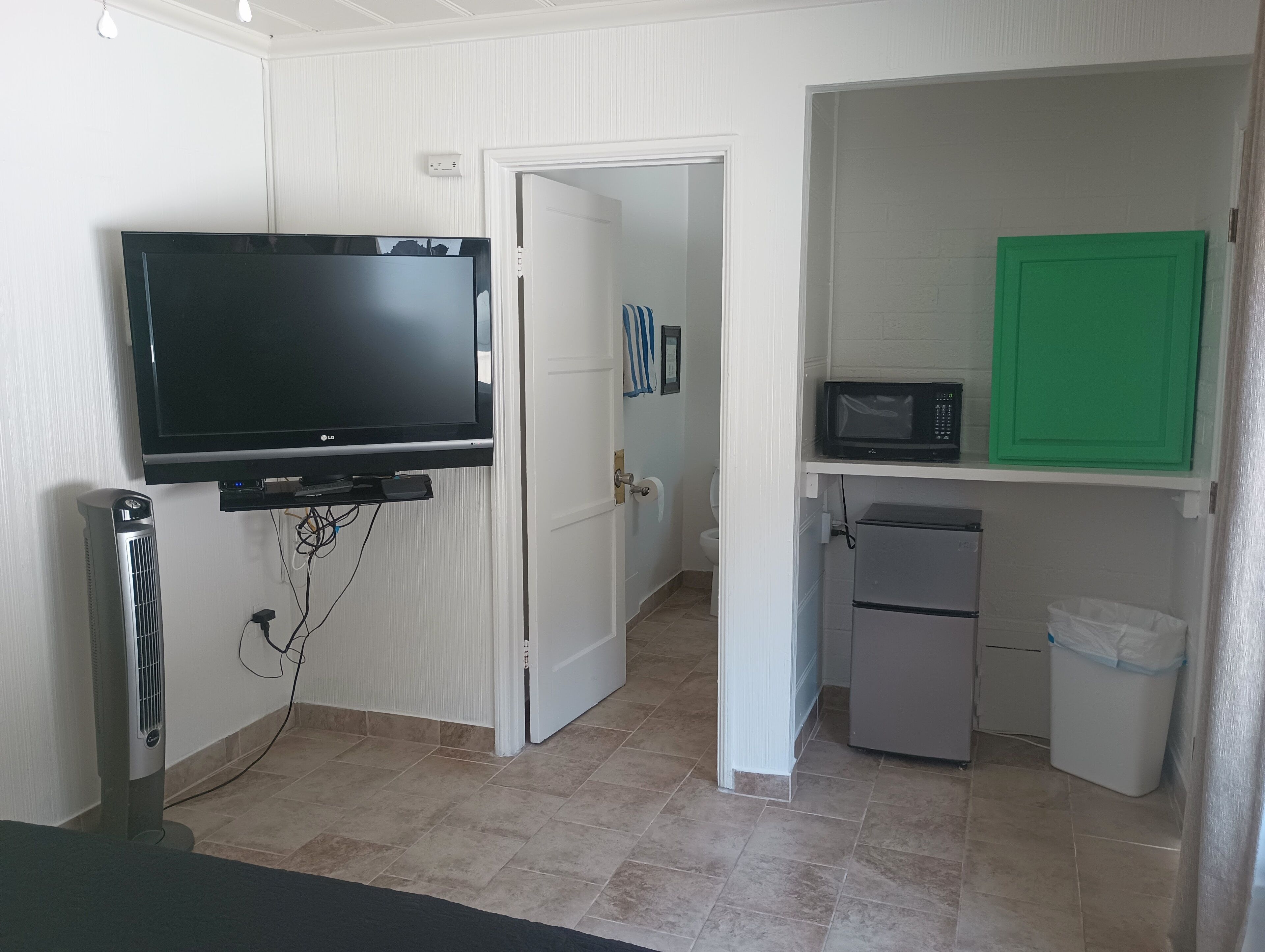 Un espacio habitable compacto con un televisor de pantalla plana montado en la pared, un refrigerador pequeño, un microondas y un cesto de basura.