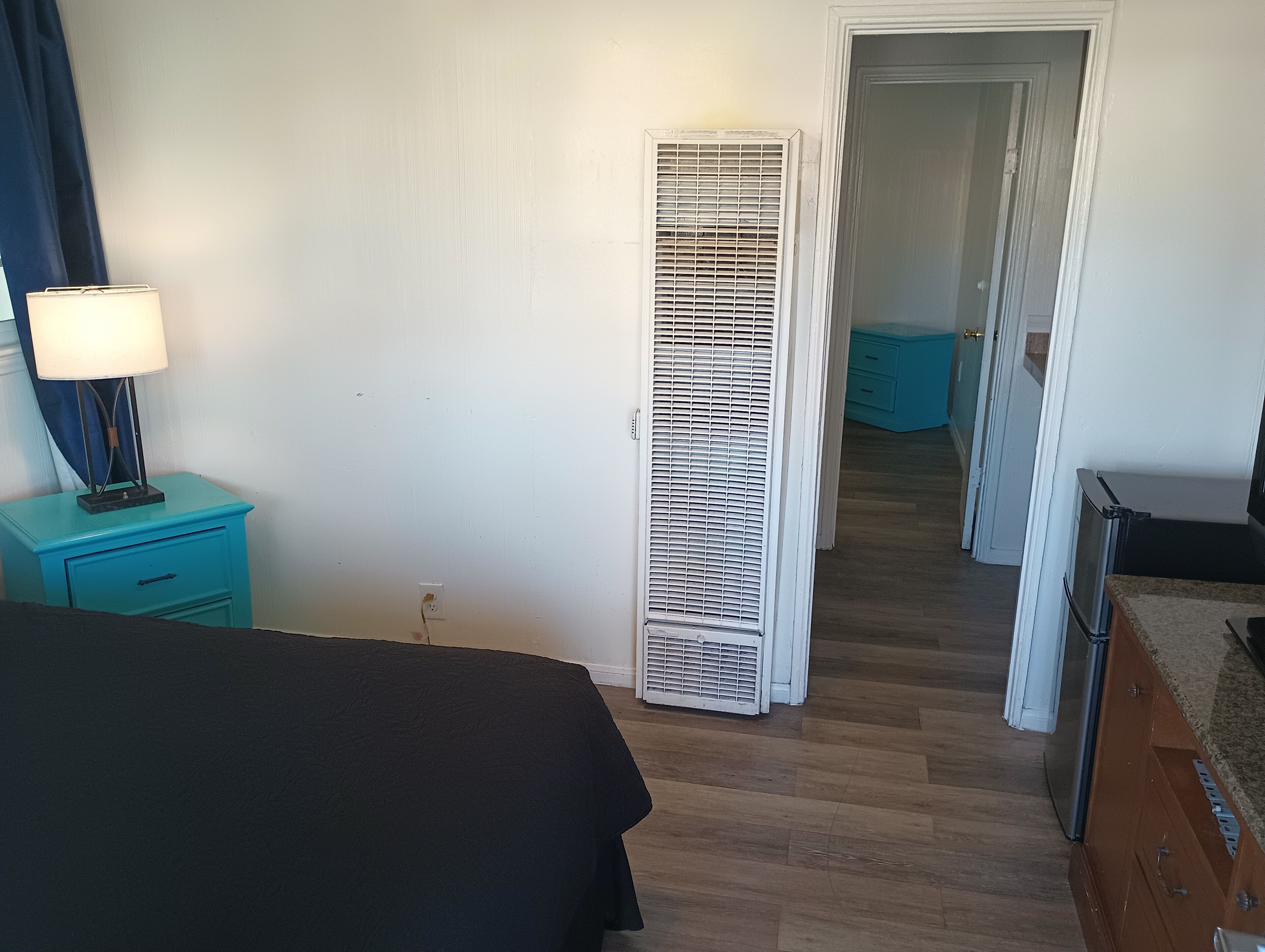 Una habitación de hotel con una cama, una mesita de noche con lámpara, un aire acondicionado alto y un aparador de madera.
