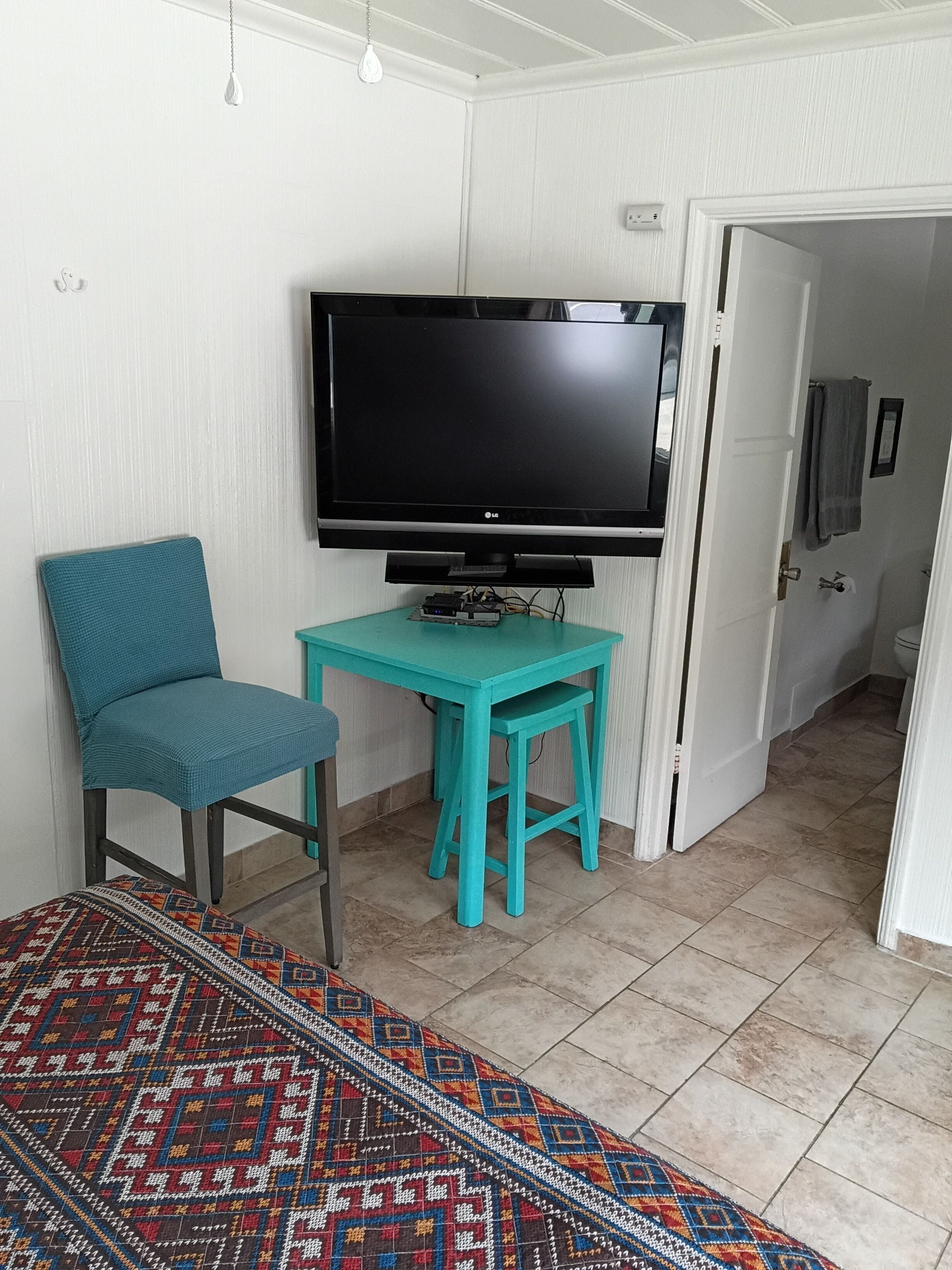 Una habitación con una televisión sobre un mueble bajo, una silla color turquesa, una mesita y una alfombra estampada.