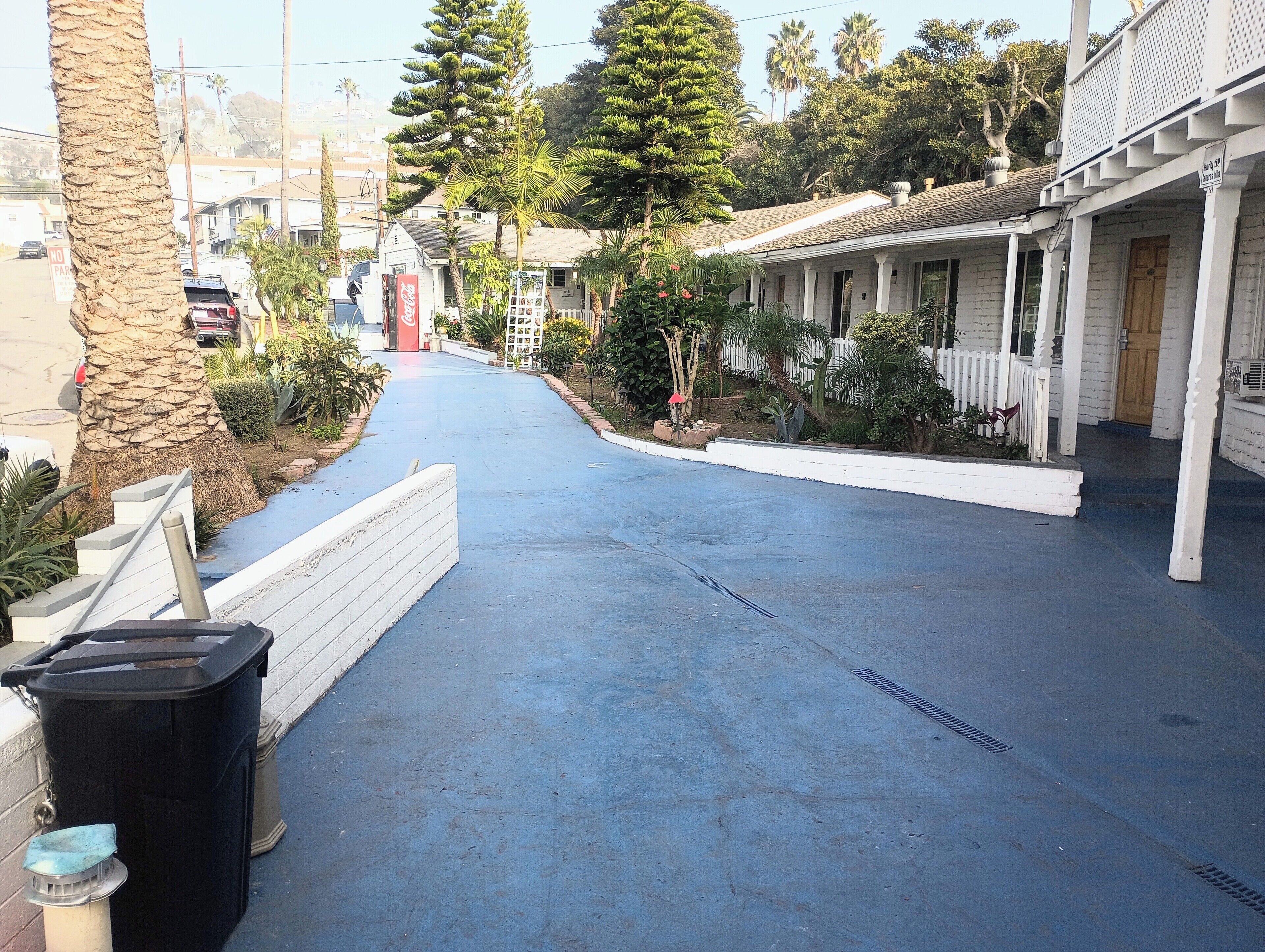 Une rue étroite avec un trottoir bleu, une poubelle, un palmier et une rangée de bâtiments aux façades blanches, sur laquelle on peut voir une enseigne Coca-Cola rouge.