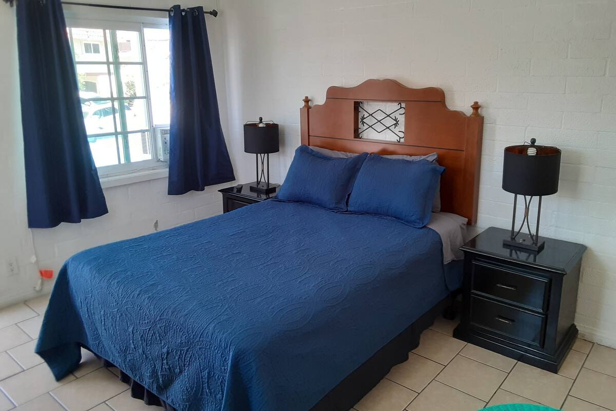 Un dormitorio con cama, dos mesitas de noche, una ventana con cortinas y una lámpara de pared.