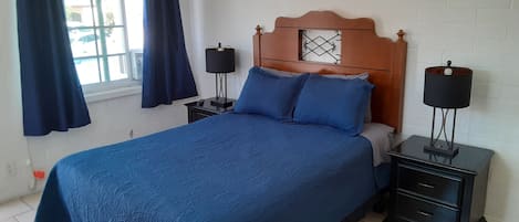Un dormitorio con cama, dos mesitas de noche, una ventana con cortinas y una lámpara de pared.