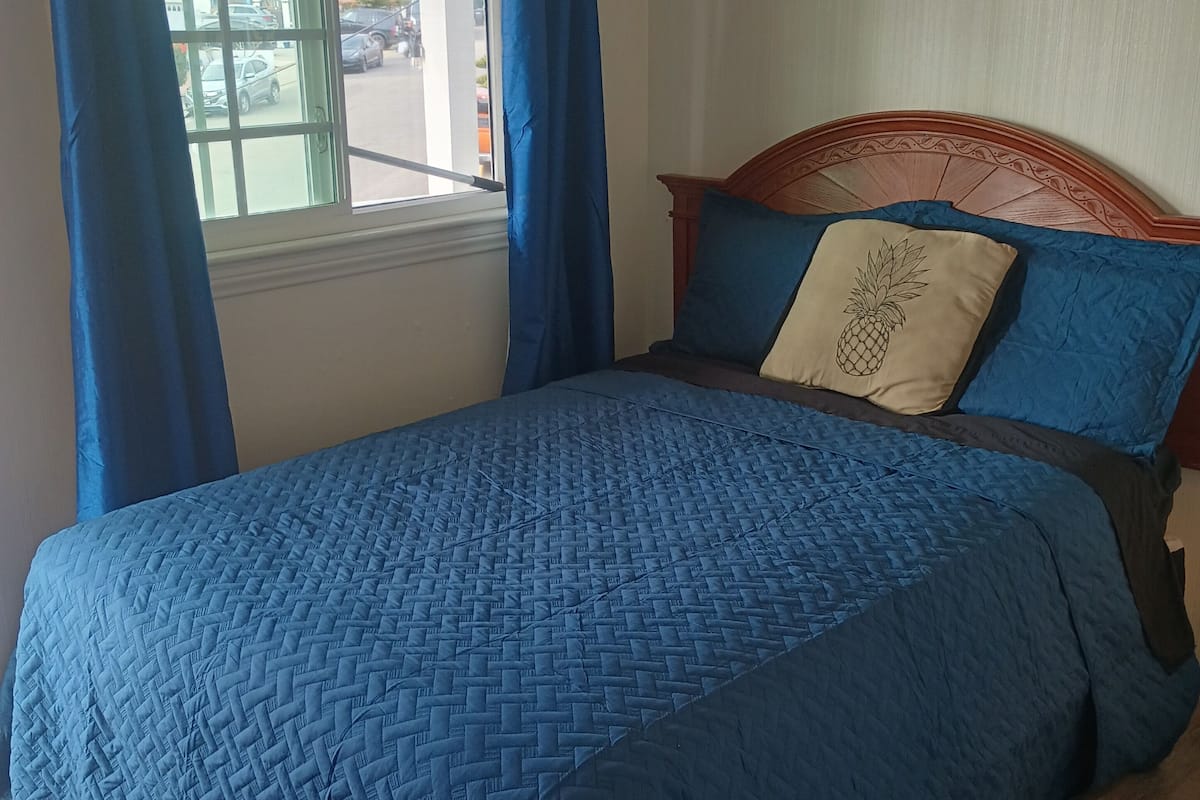 Una cama con colcha azul, cabecera de madera y ventana con cortinas azules.
