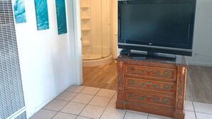 Une pièce avec un téléviseur posé sur un meuble en bois, une porte de douche en verre et trois œuvres d’art accrochées au mur.
