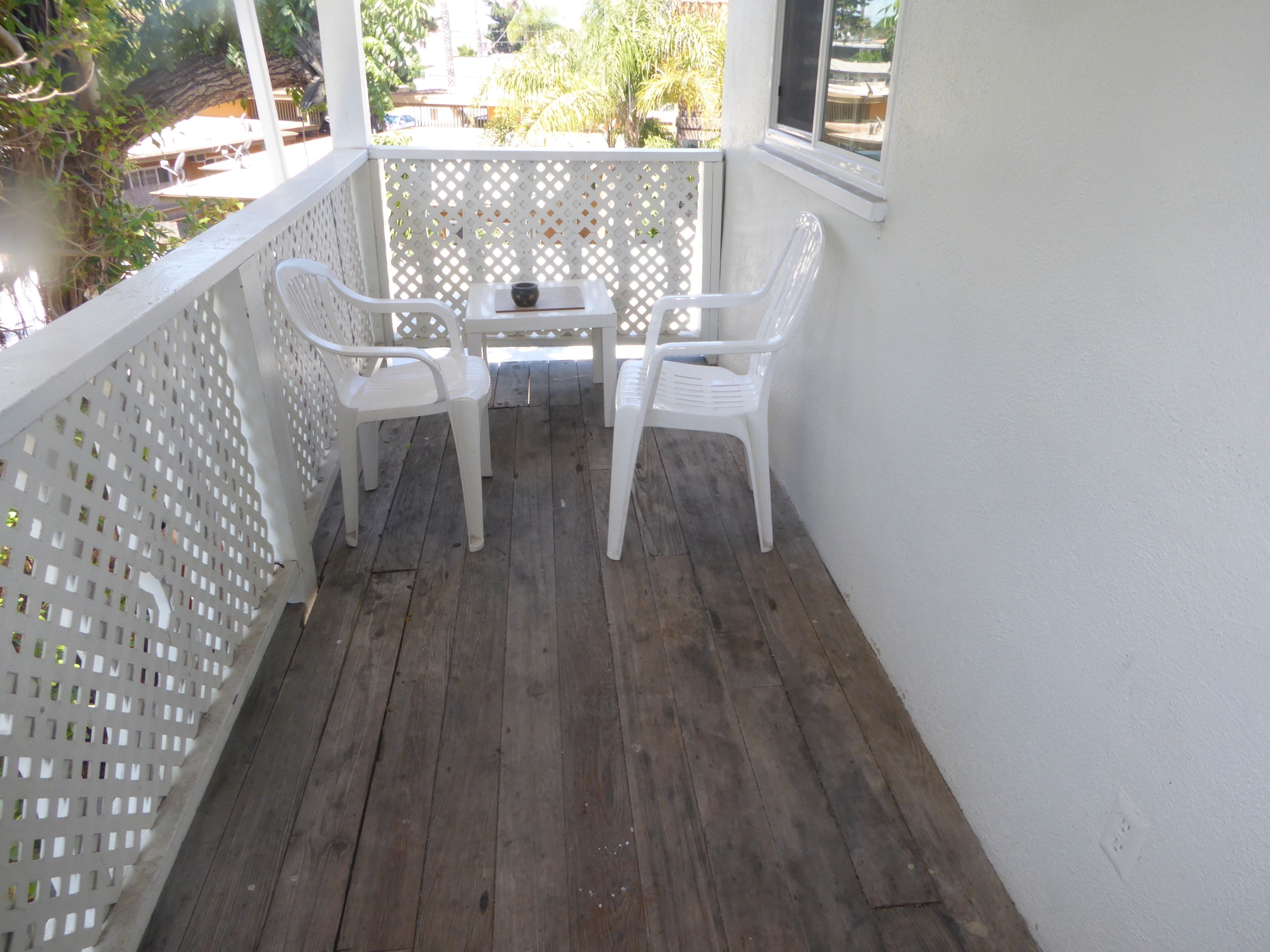 Een balkon met twee witte plastic stoelen en een kleine tafel, houten vloeren en een wit lattenhek.
