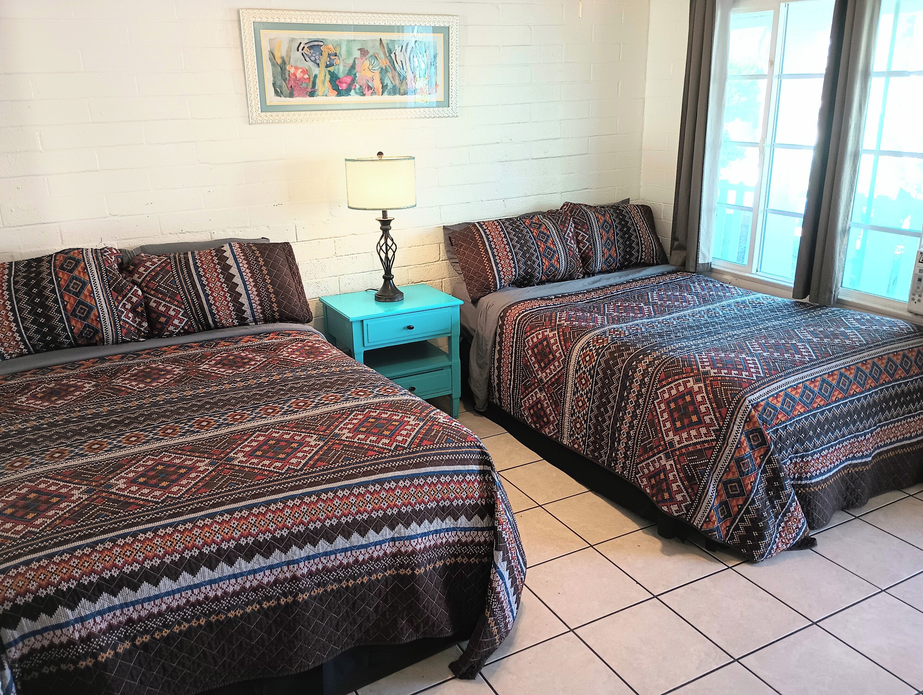 Una habitación de hotel con dos camas, una mesita con lámpara, un cuadro en la pared y una ventana con cortinas.