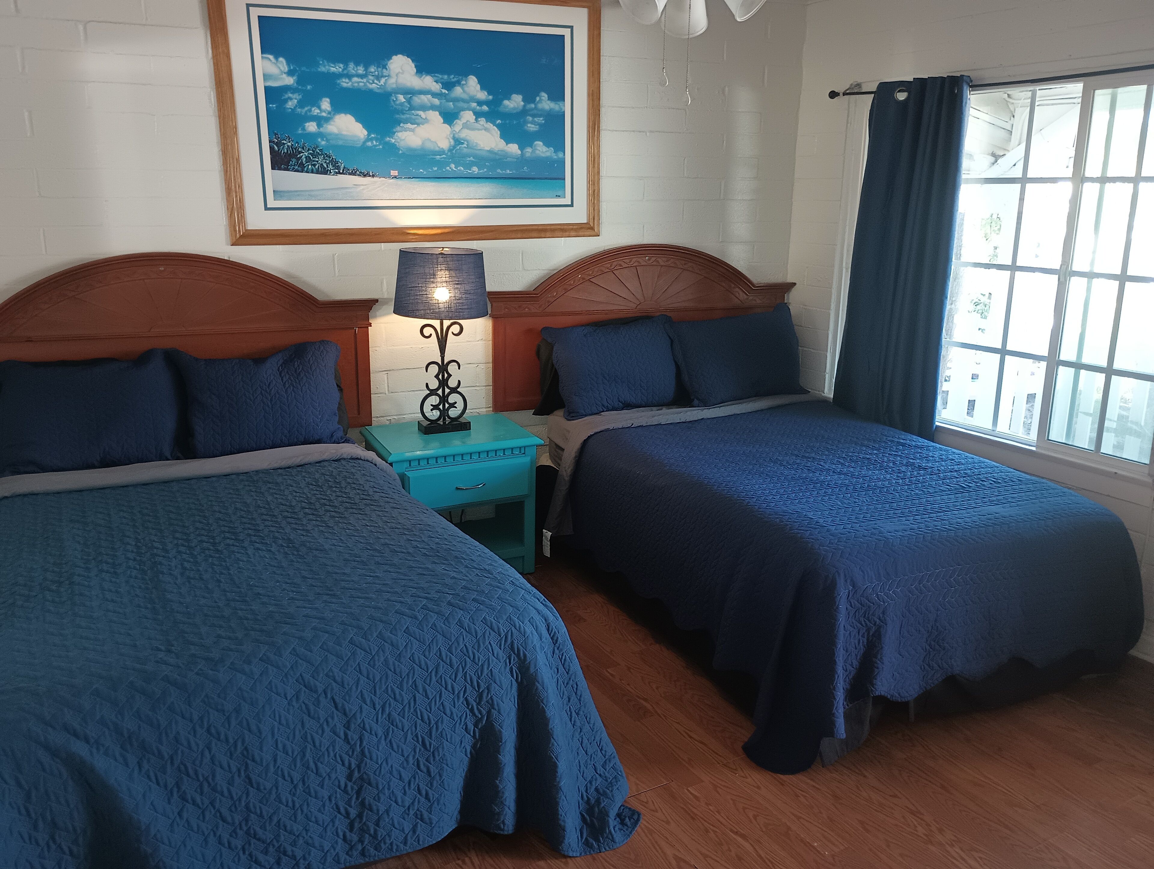 A room with two beds, a nightstand with a lamp, a framed picture on the wall, and a window with curtains.