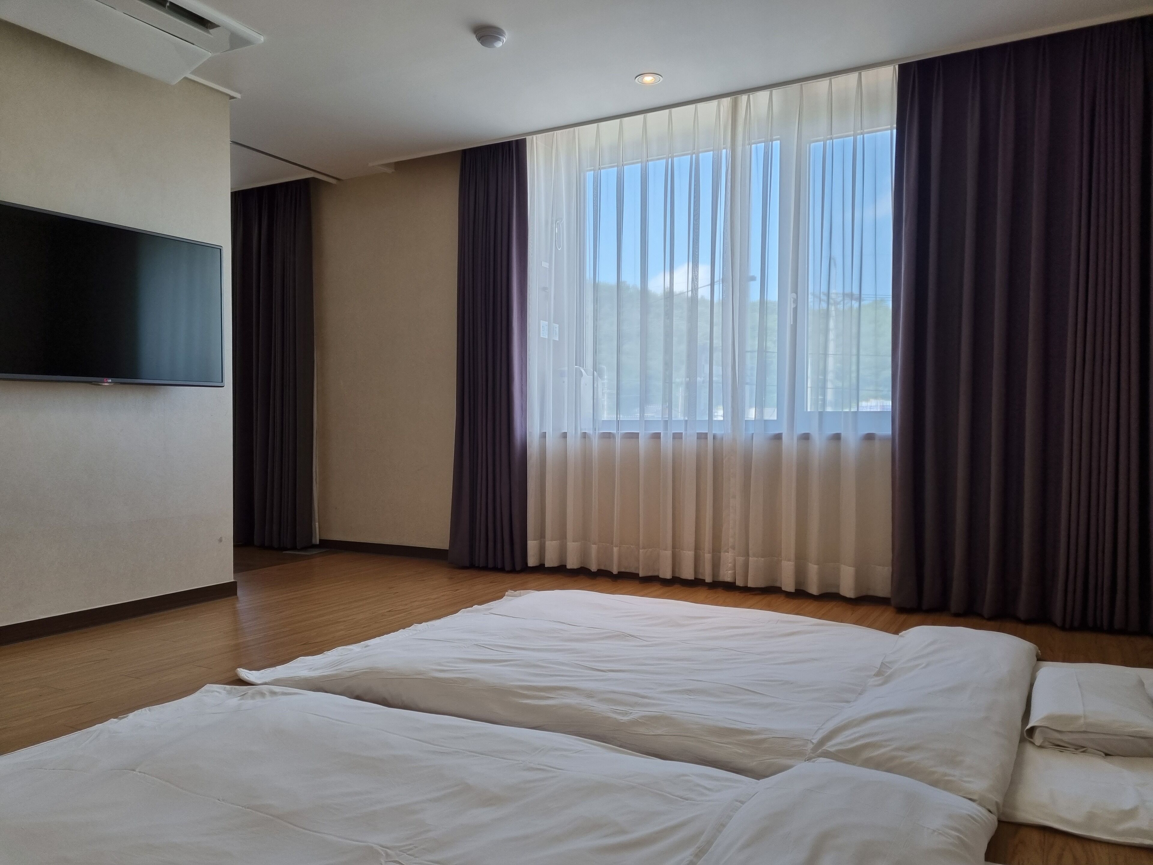 Habitación de hotel con dos camas, un televisor de pantalla plana montado en la pared y ventanas amplias con cortinas translúcidas y opacas.