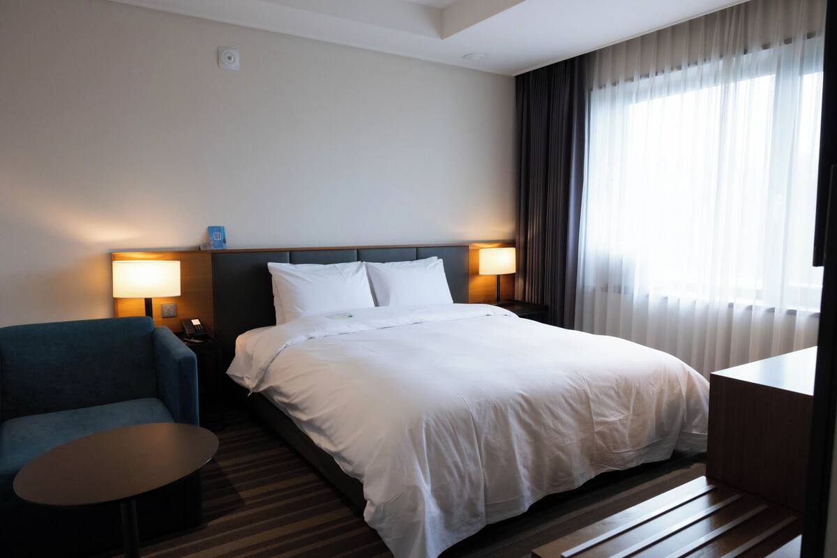 A hotel room with a bed, two bedside lamps, a small table, and a chair.